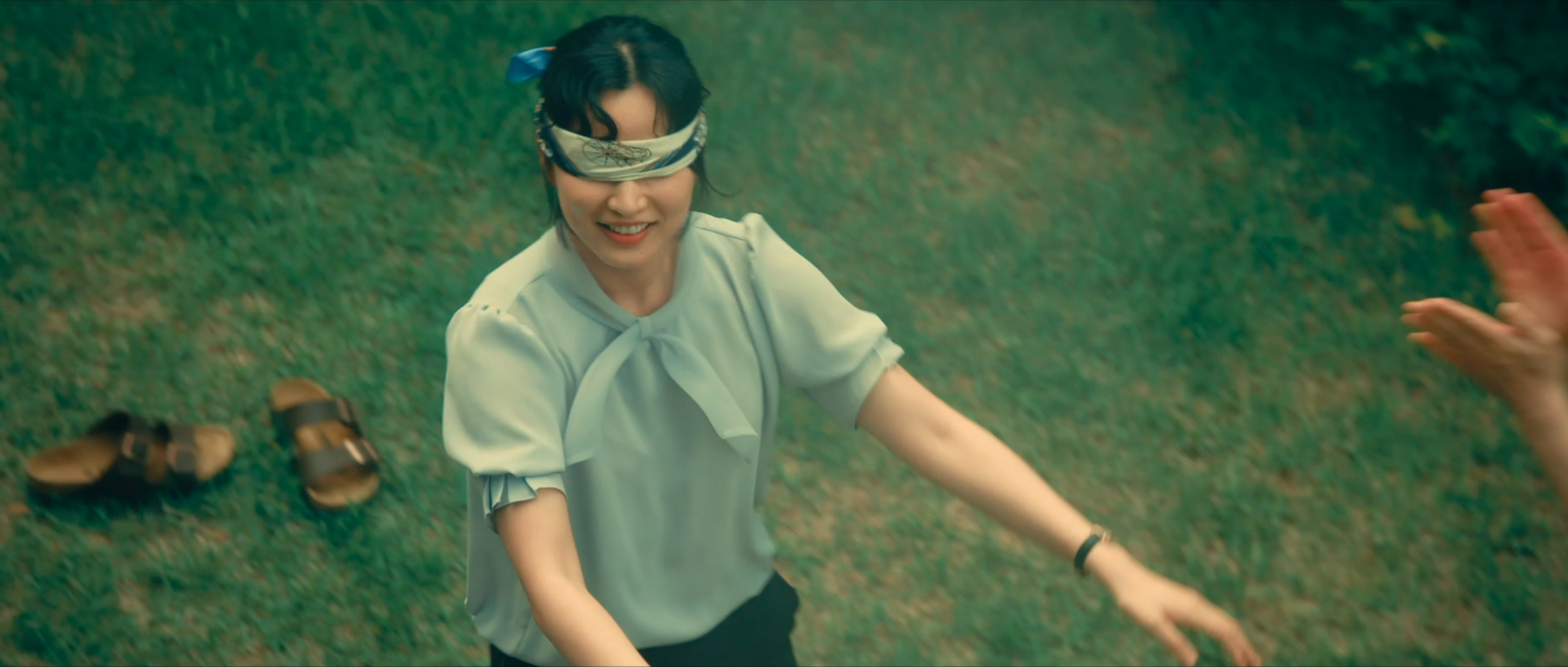 A blindfolded woman smiling and reaching forward on a grassy field as another person claps nearby.