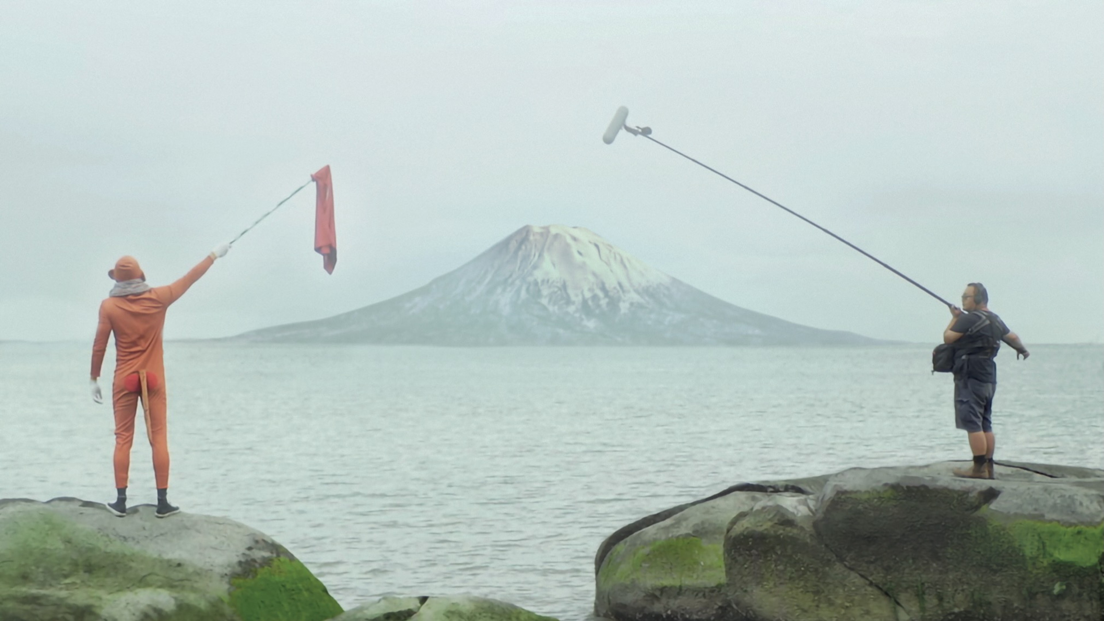 A person in an orange outfit waving a flag from a rocky shoreline while another person holds a boom microphone, with a snow-capped mountain across the water.