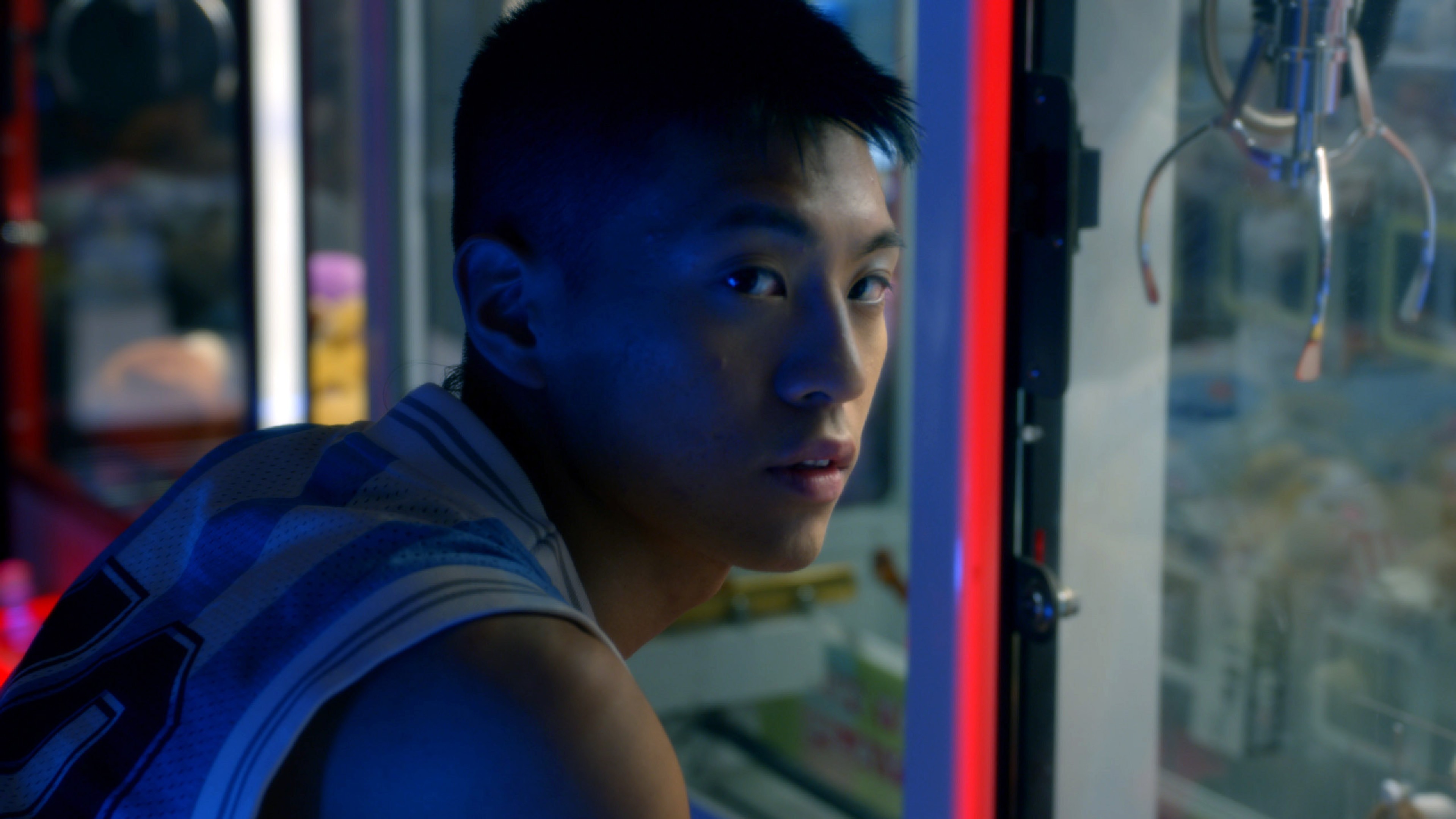 A young man looking back over his shoulder beside a brightly lit arcade claw machine.