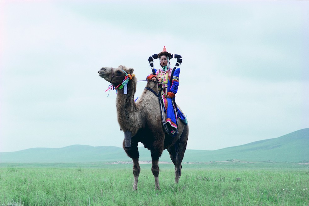 A person in colourful traditional dress riding a camel across a wide Mongolian grassland.
