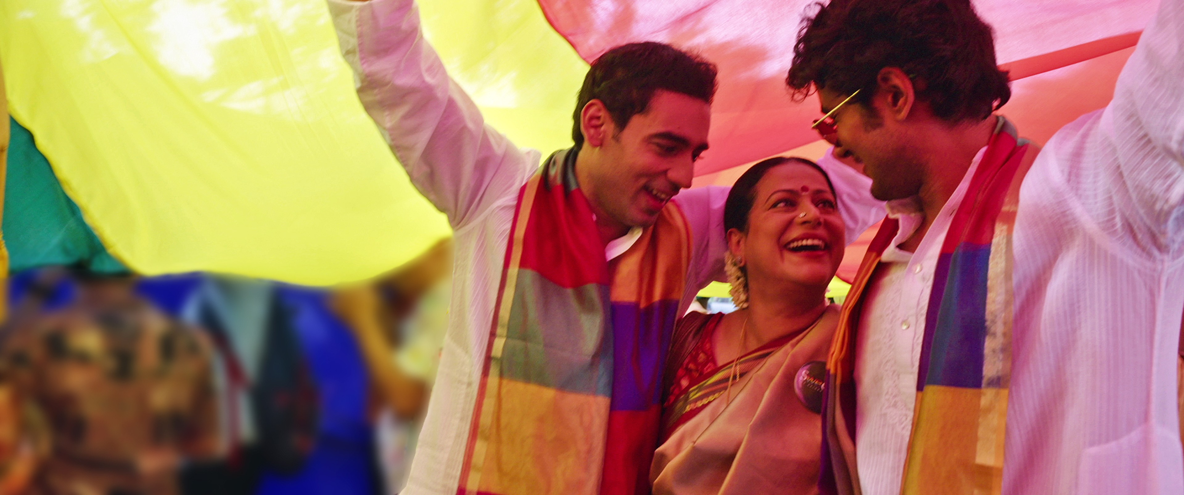 Three people smiling together beneath rainbow-coloured fabric, two wearing rainbow scarves.