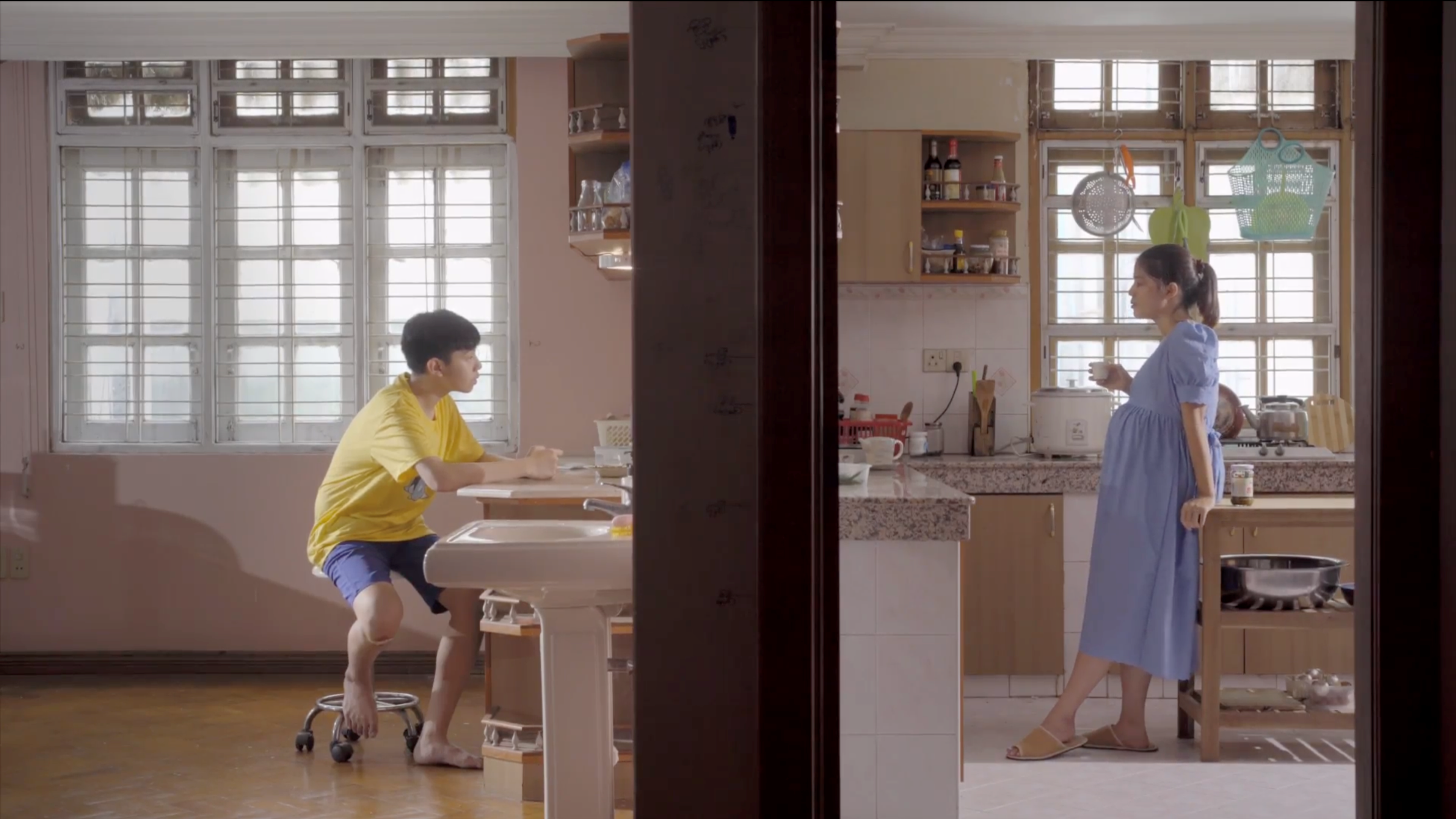 A person seated at a dining table while another stands in a bright kitchen across the room.