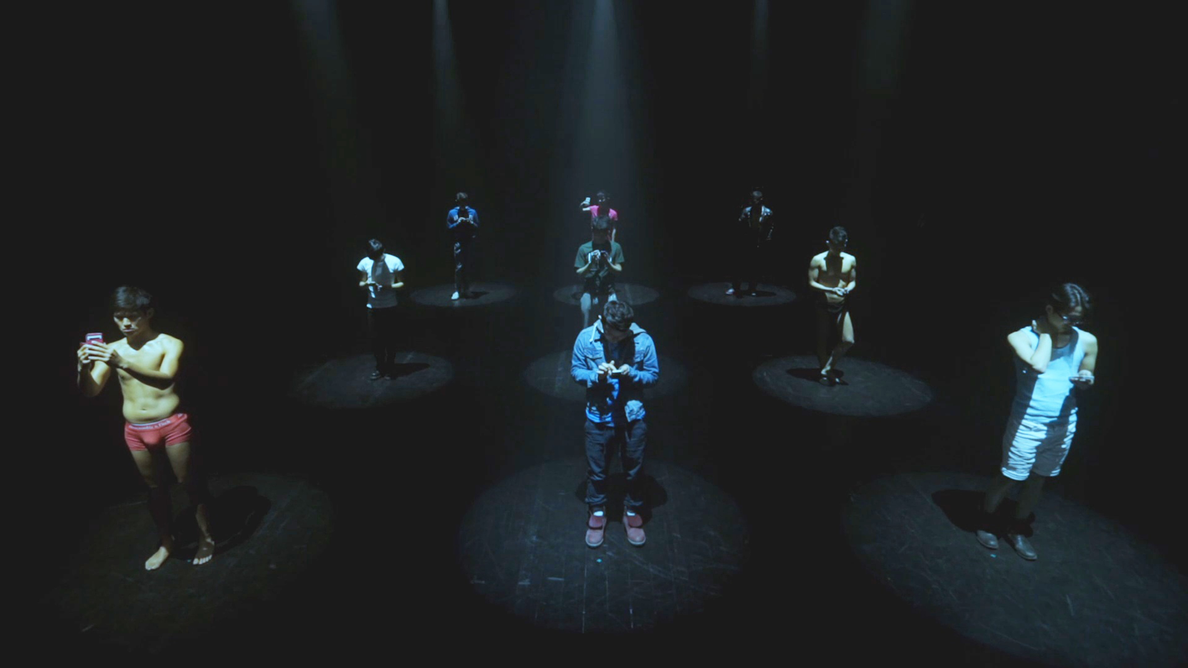 nine people each holding a phone and standing in a spotlight.