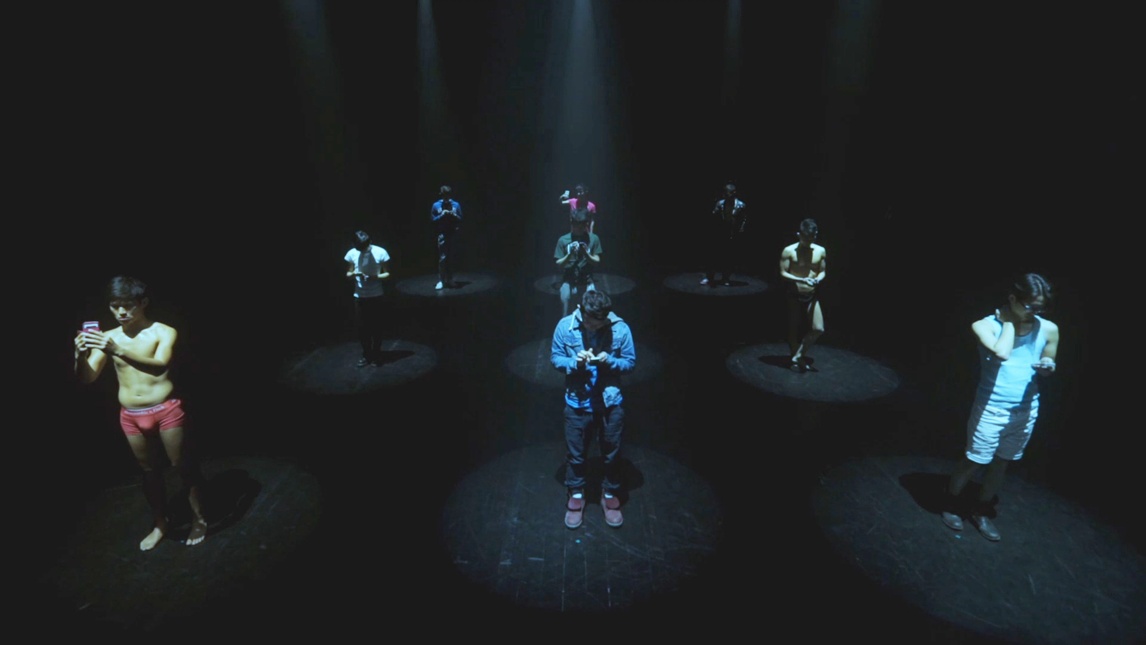 nine people each holding a phone and standing in a spotlight.