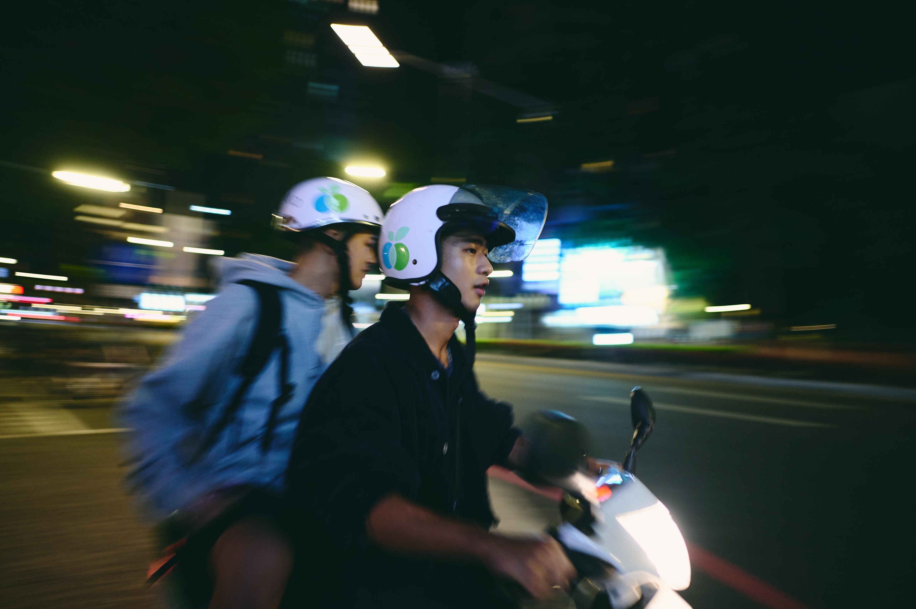 Two people wearing helmets riding together on a scooter through a city street at night.