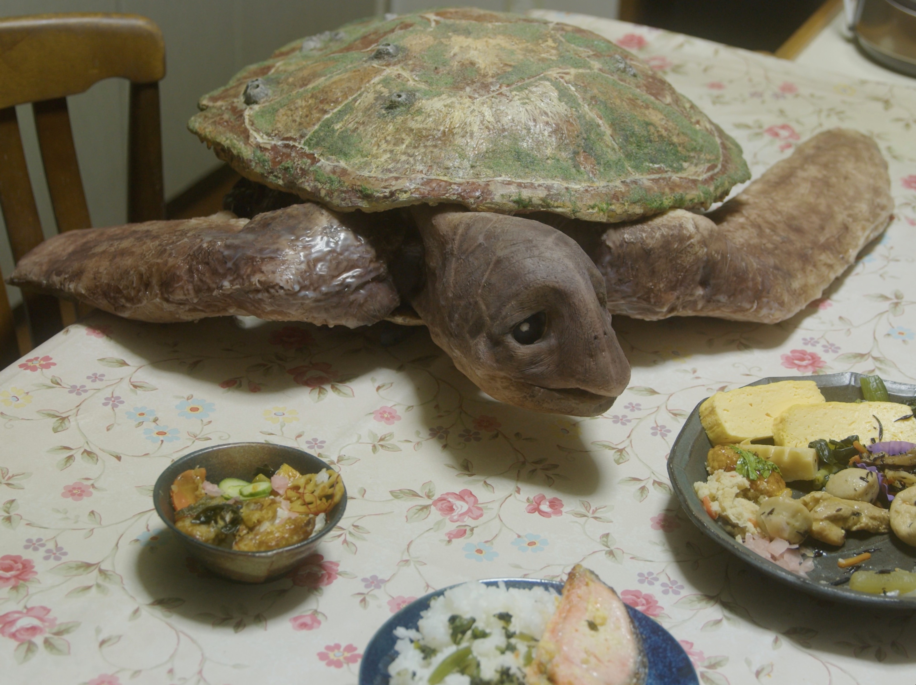 A large turtle sculpture rests on a dining table among plates and bowls of food.