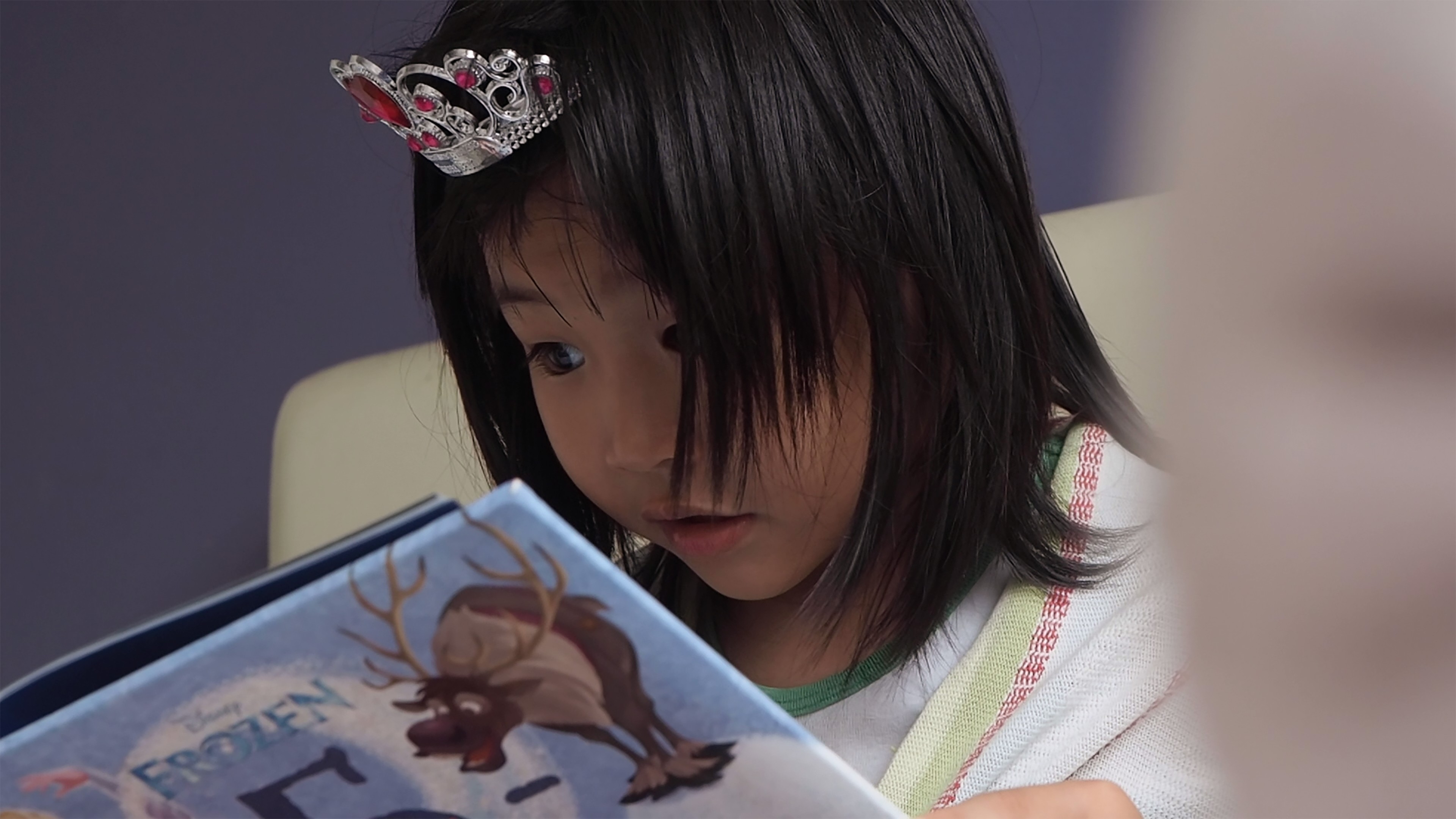 A young child wearing a small tiara reading a picture book.
