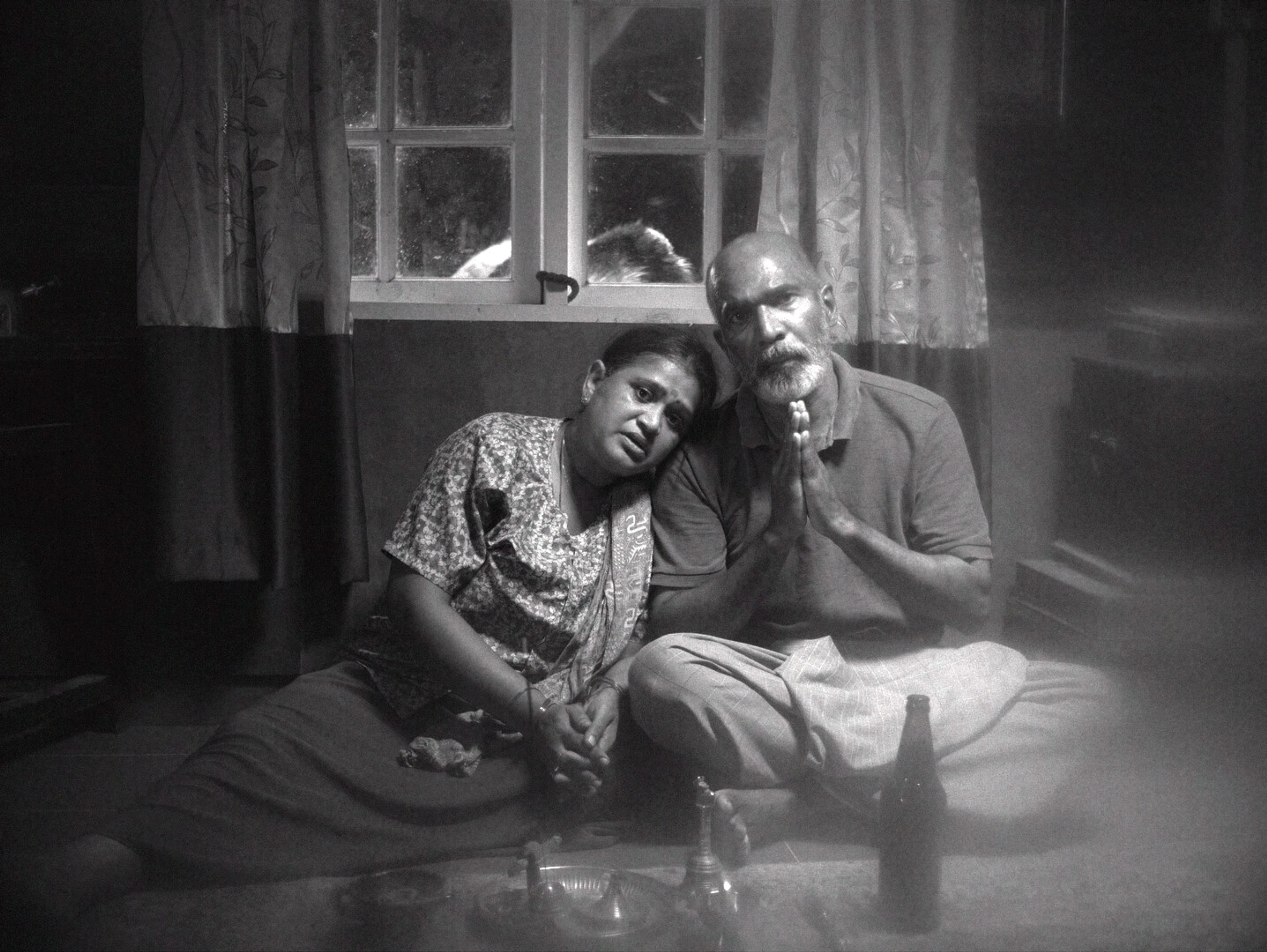 Two people sit under a window with their hands clasped in prayer.