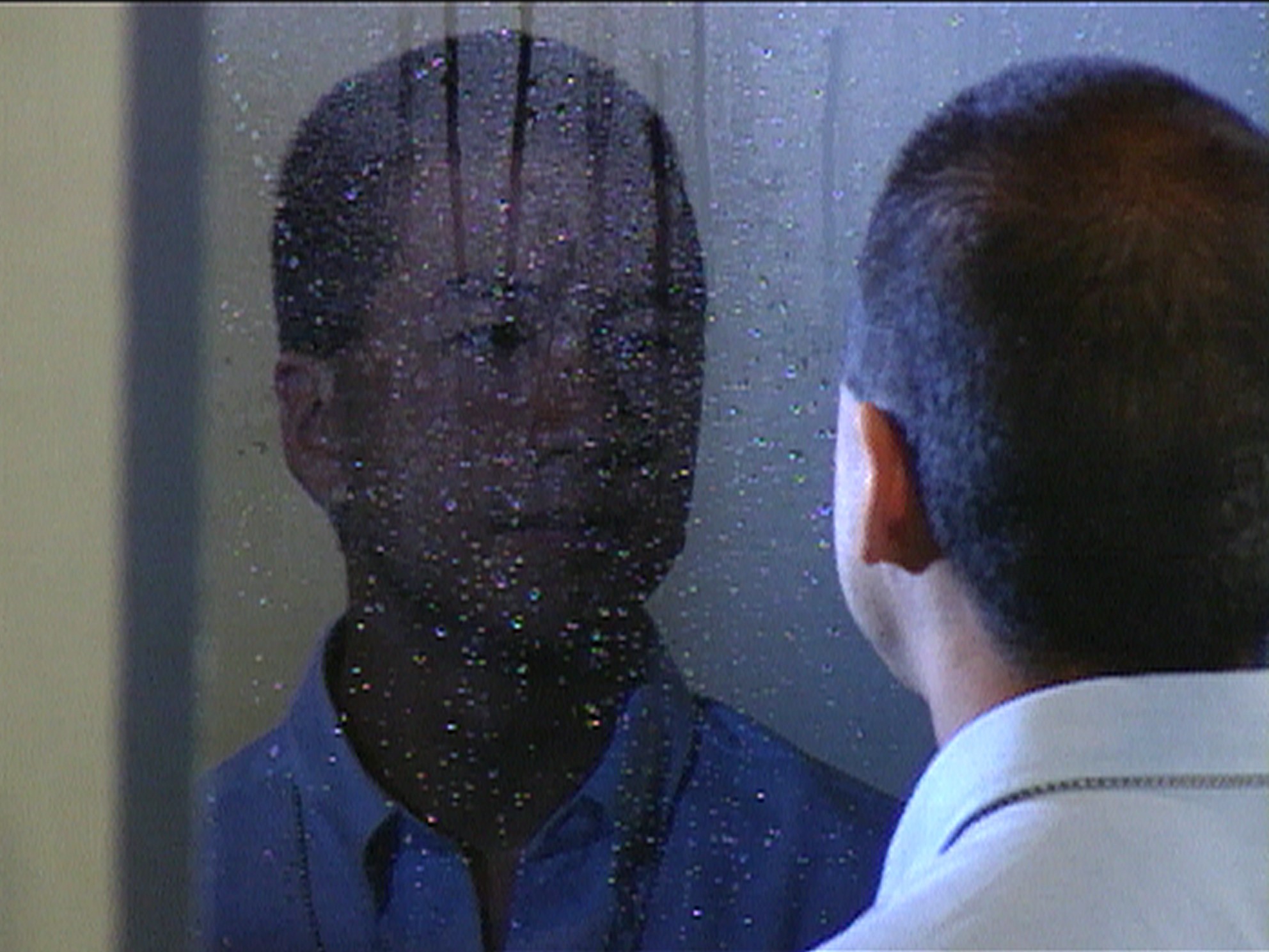 A person looking at their reflection in a rain-speckled mirror, seen from behind in a dimly lit room.