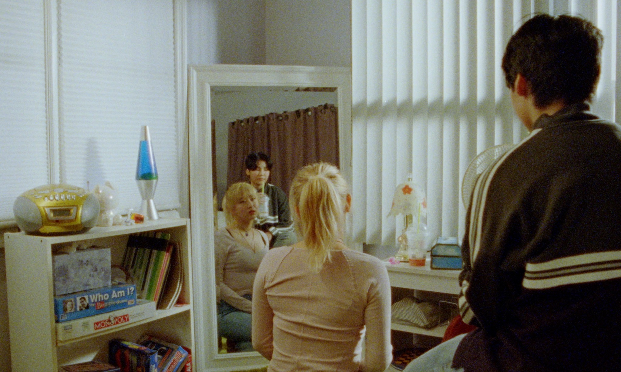 Three people gathered in a bedroom, with one seated facing a mirror and another standing nearby.