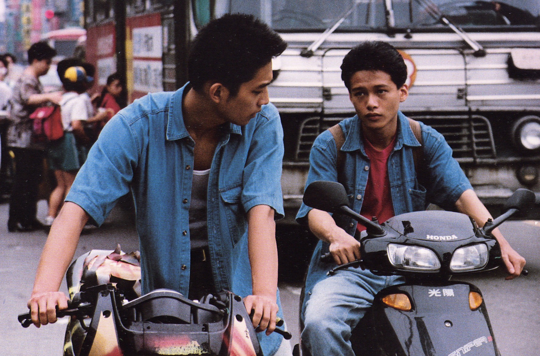 Two young men sitting on scooters in a busy street, looking toward each other.