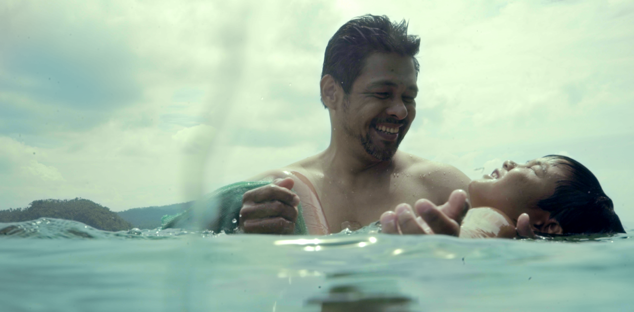 Two people smiling together while swimming in open water, one supporting the other as they float.