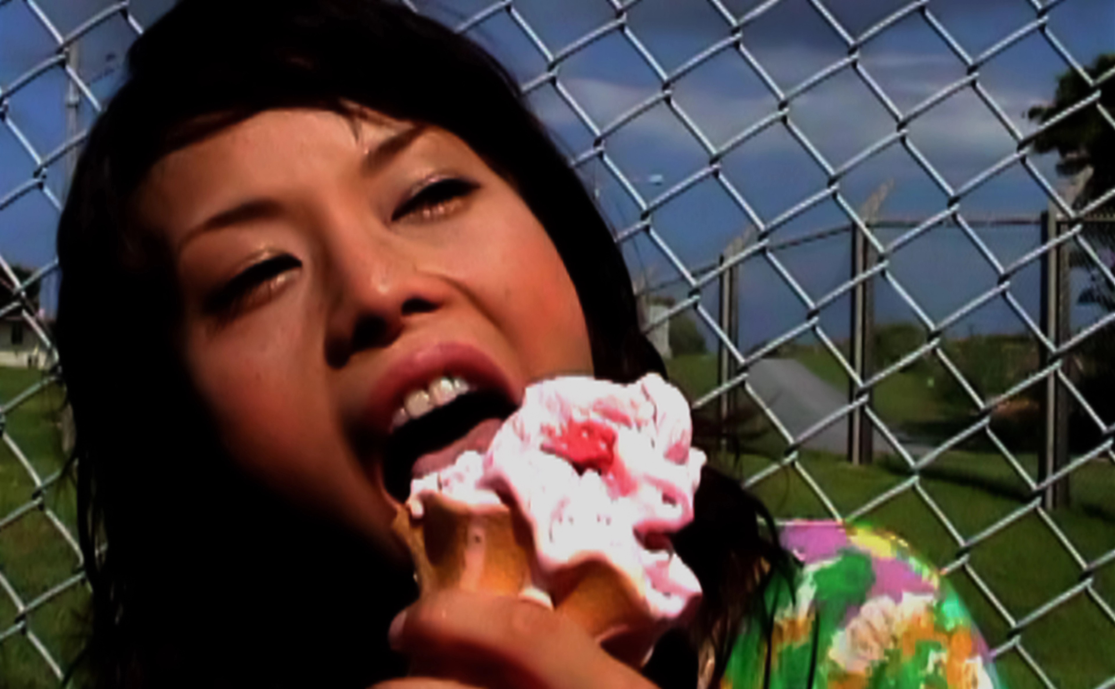 A woman eats a melting ice cream against a chainlink fence.