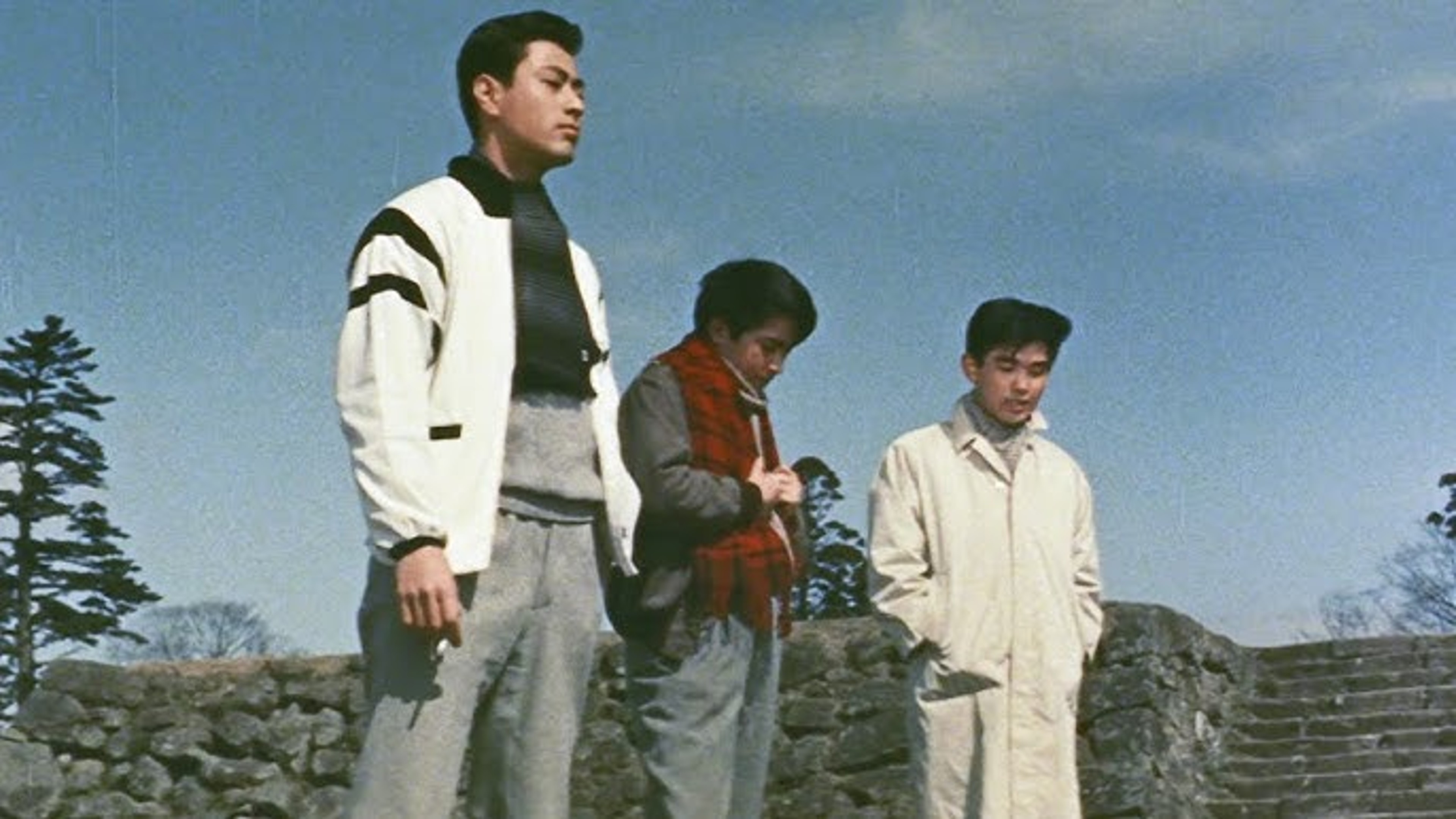 Three men standing outdoors beside a stone wall under a blue sky.
