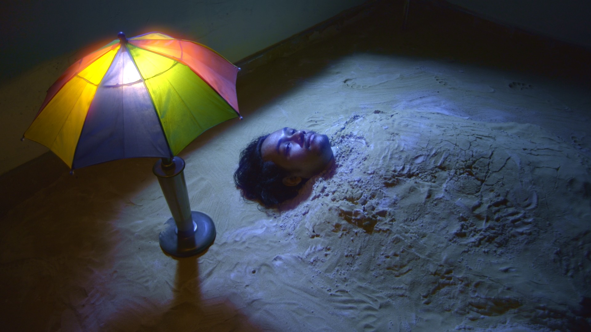 a man buried up to his neck in the sand is illuminated by a colourful lamp.