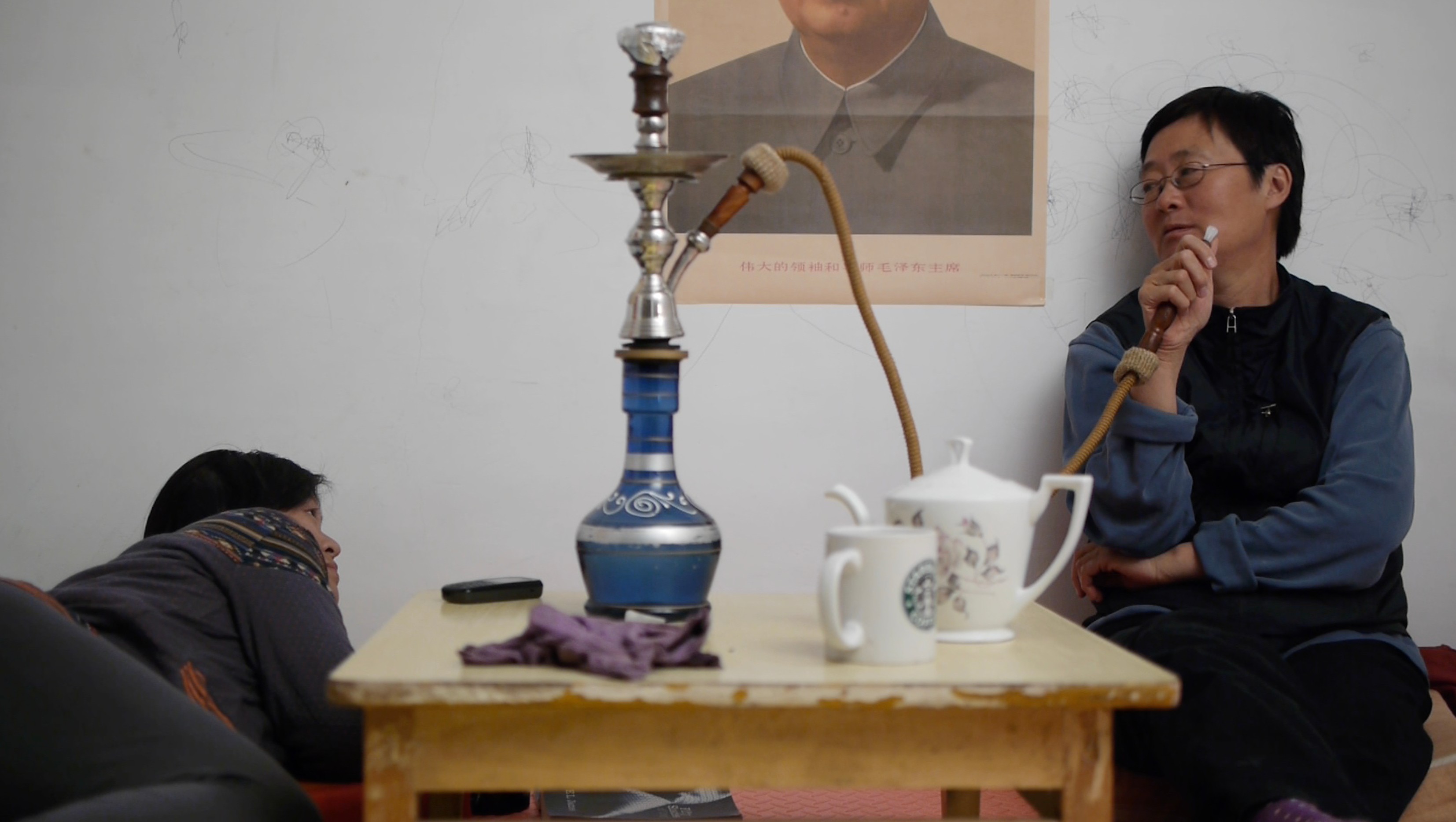 Two people relaxing in a small room beside a table with a hookah, teapot and mugs.