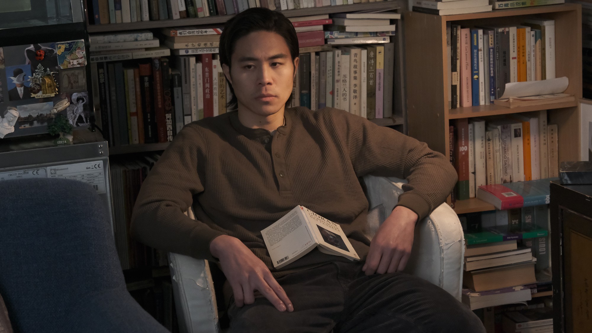 A man seated in an armchair holding an open book, surrounded by shelves filled with books.
