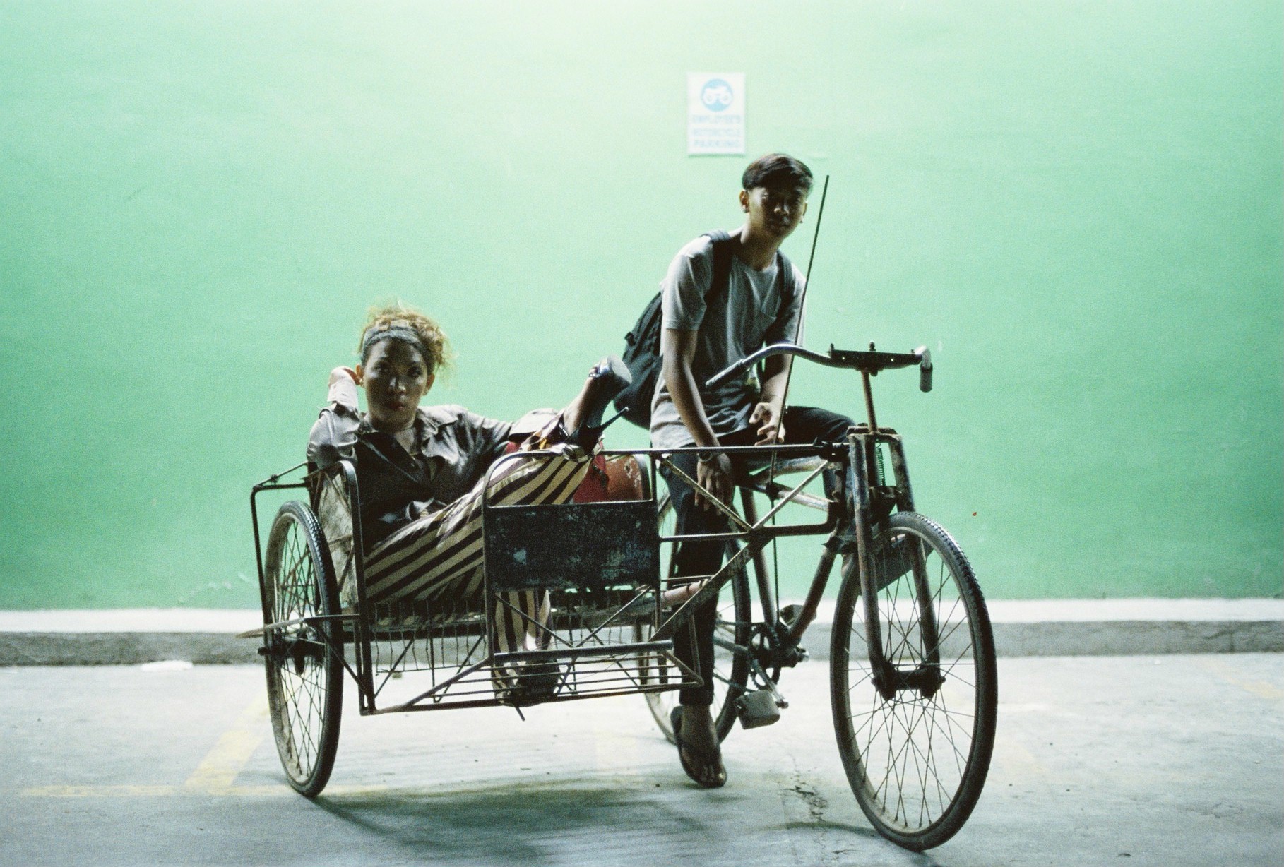 Two people riding a three-wheeled cycle together against a bright green wall.