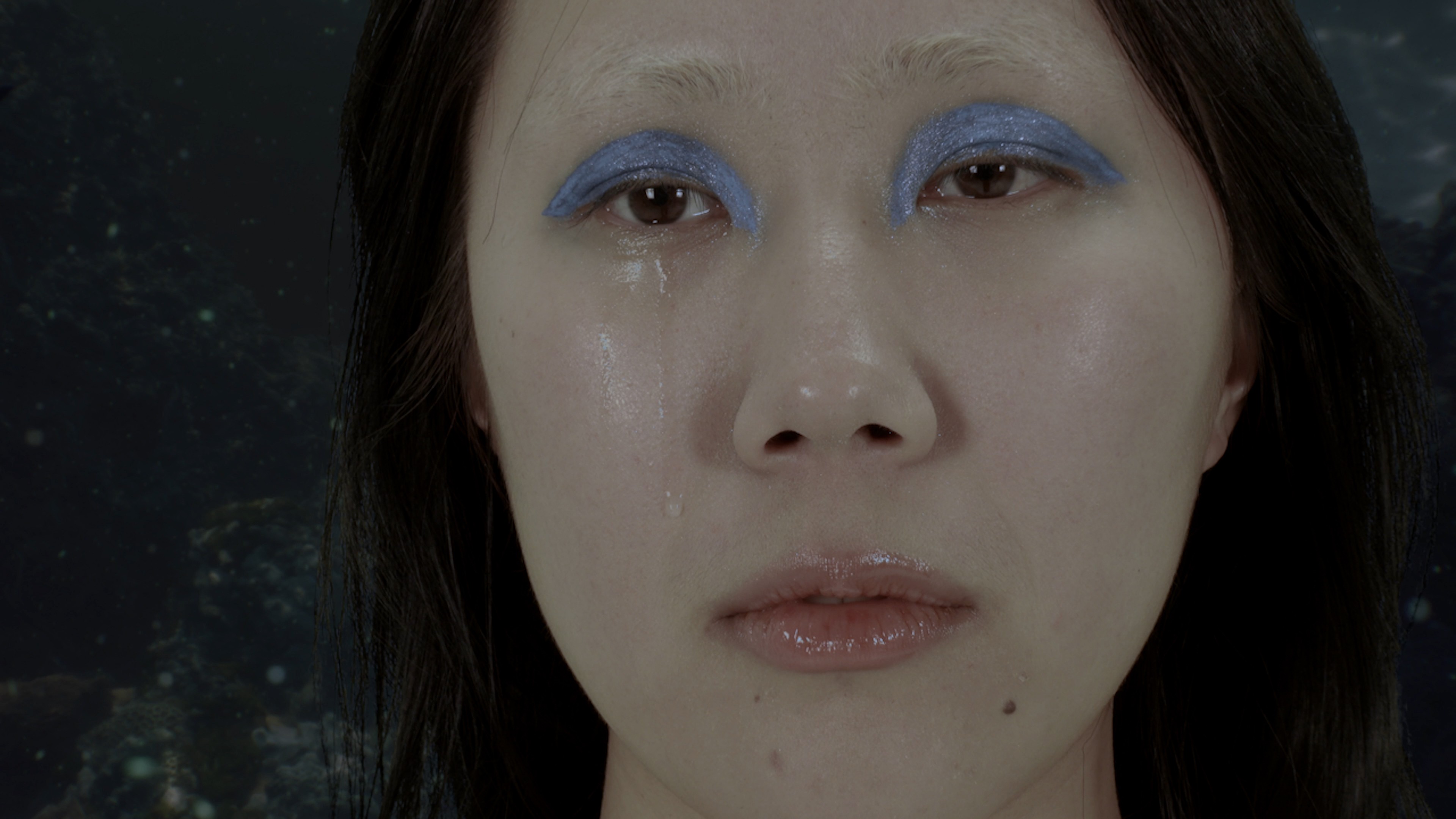 A close-up portrait of a woman with shimmering blue eye makeup and tears running down her face against a dark background.