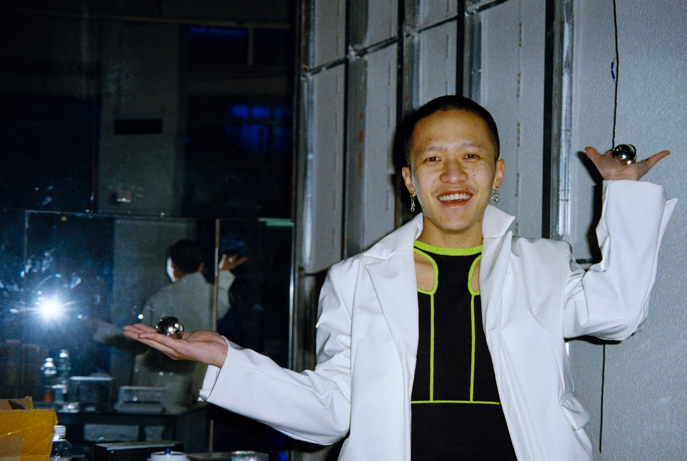 A smiling person in a white jacket posing with arms raised, holding small metallic spheres in each hand.
