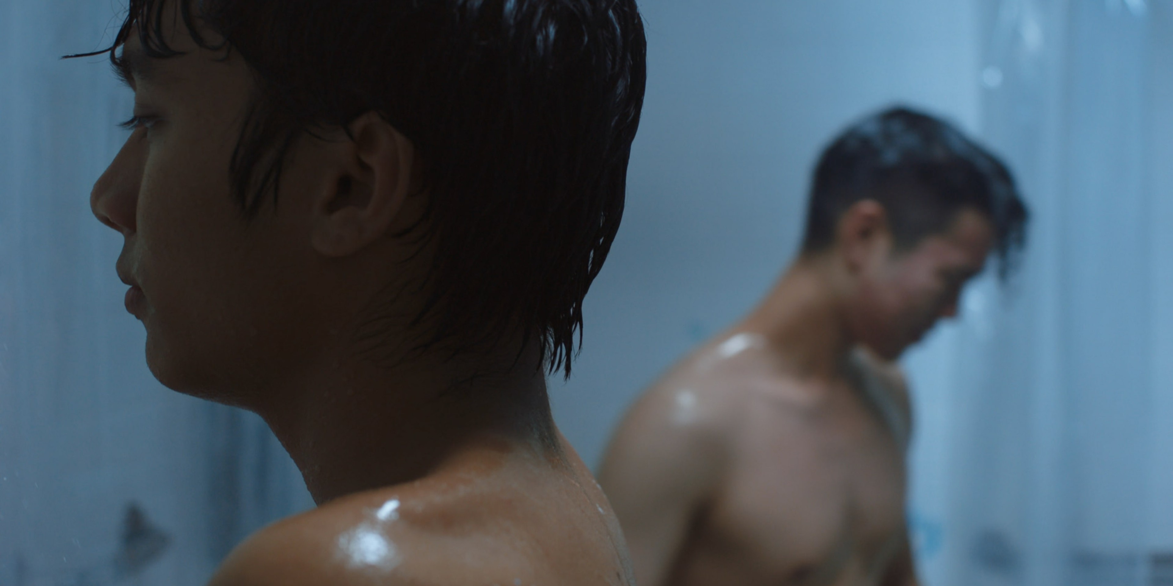 Two shirtless men standing apart in a steamy blue-lit shower room.