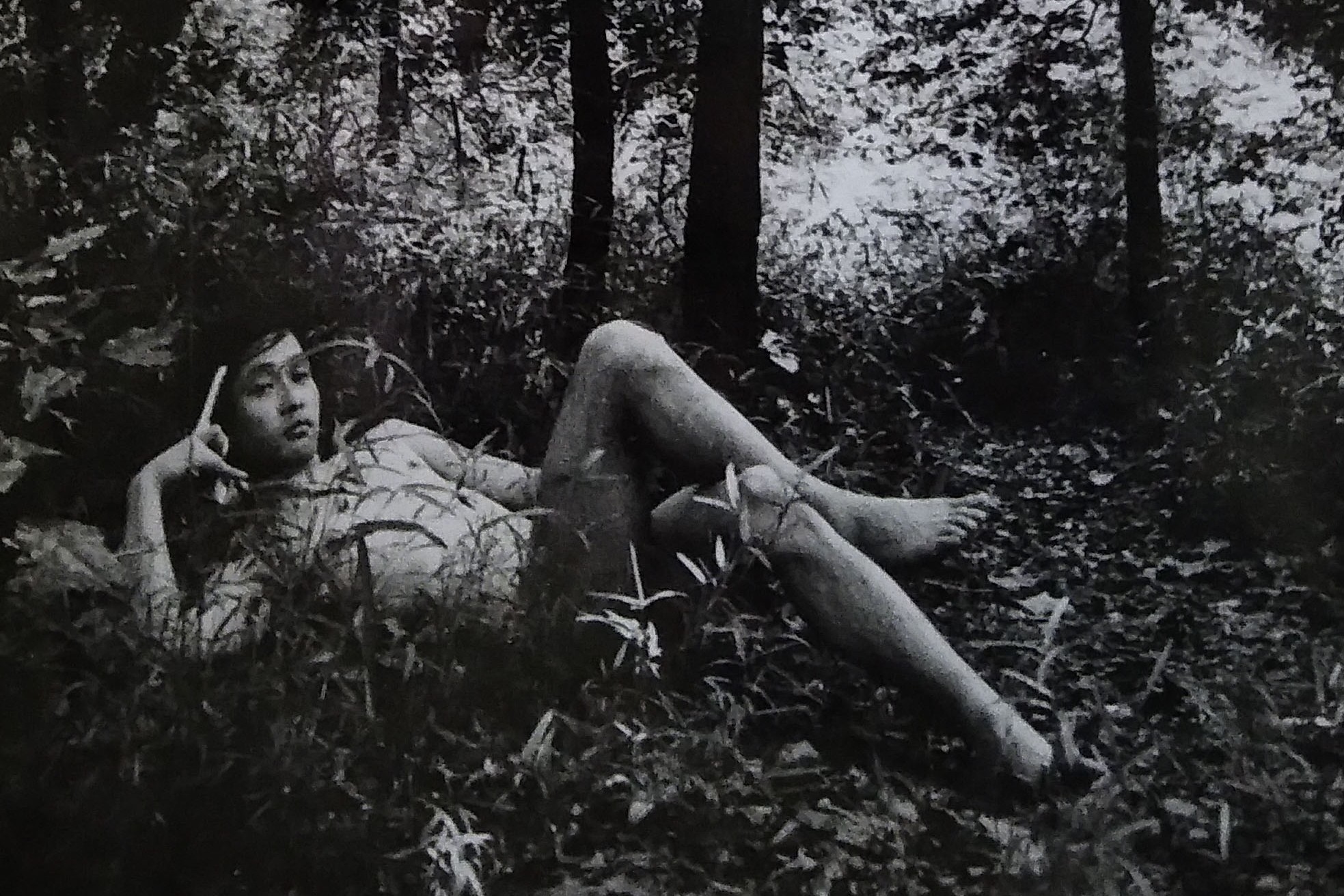 A person reclining barefoot among dense vegetation in a black-and-white forest scene.