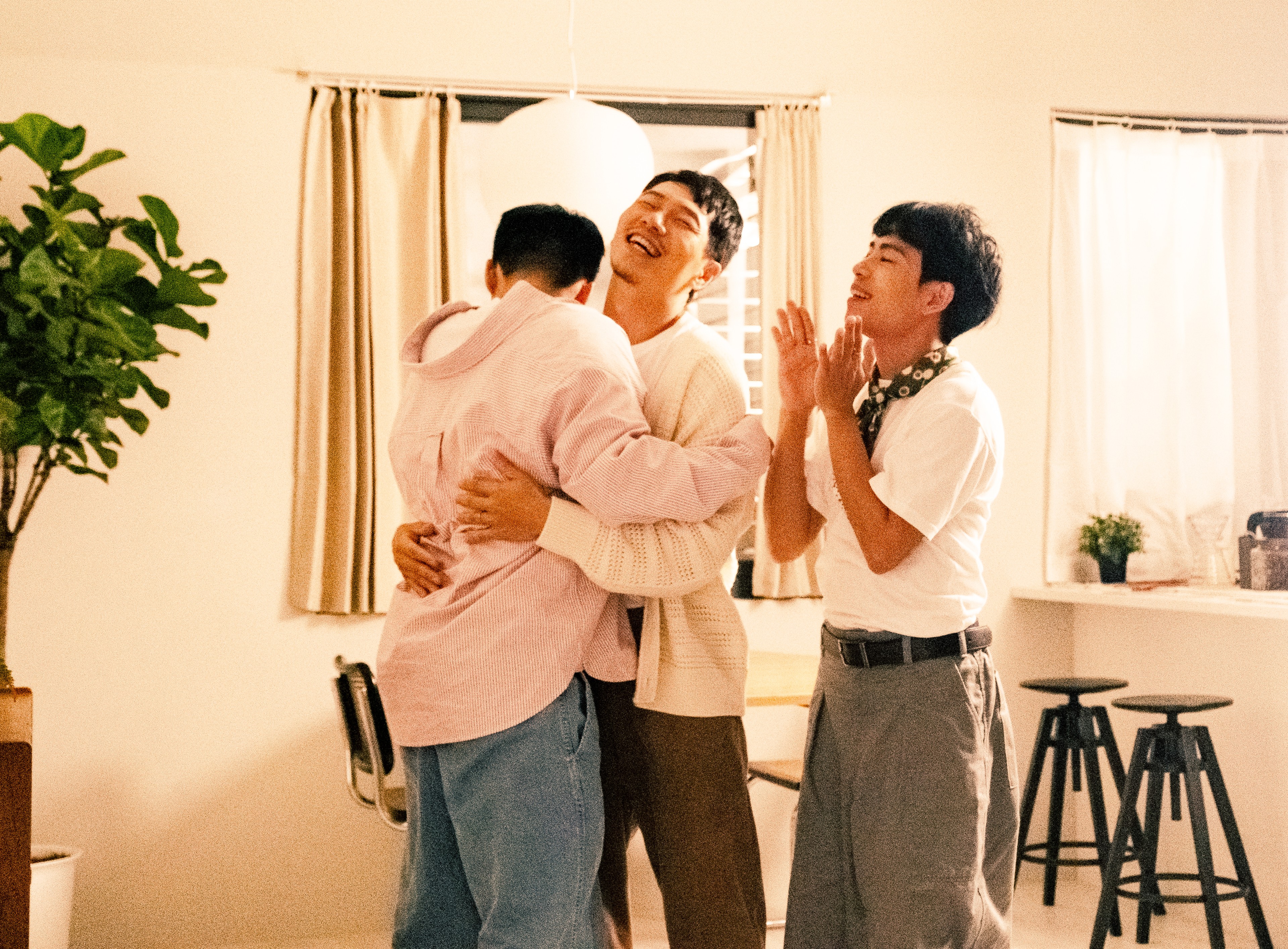 Three men celebrating together indoors, with two embracing and another clapping beside them.