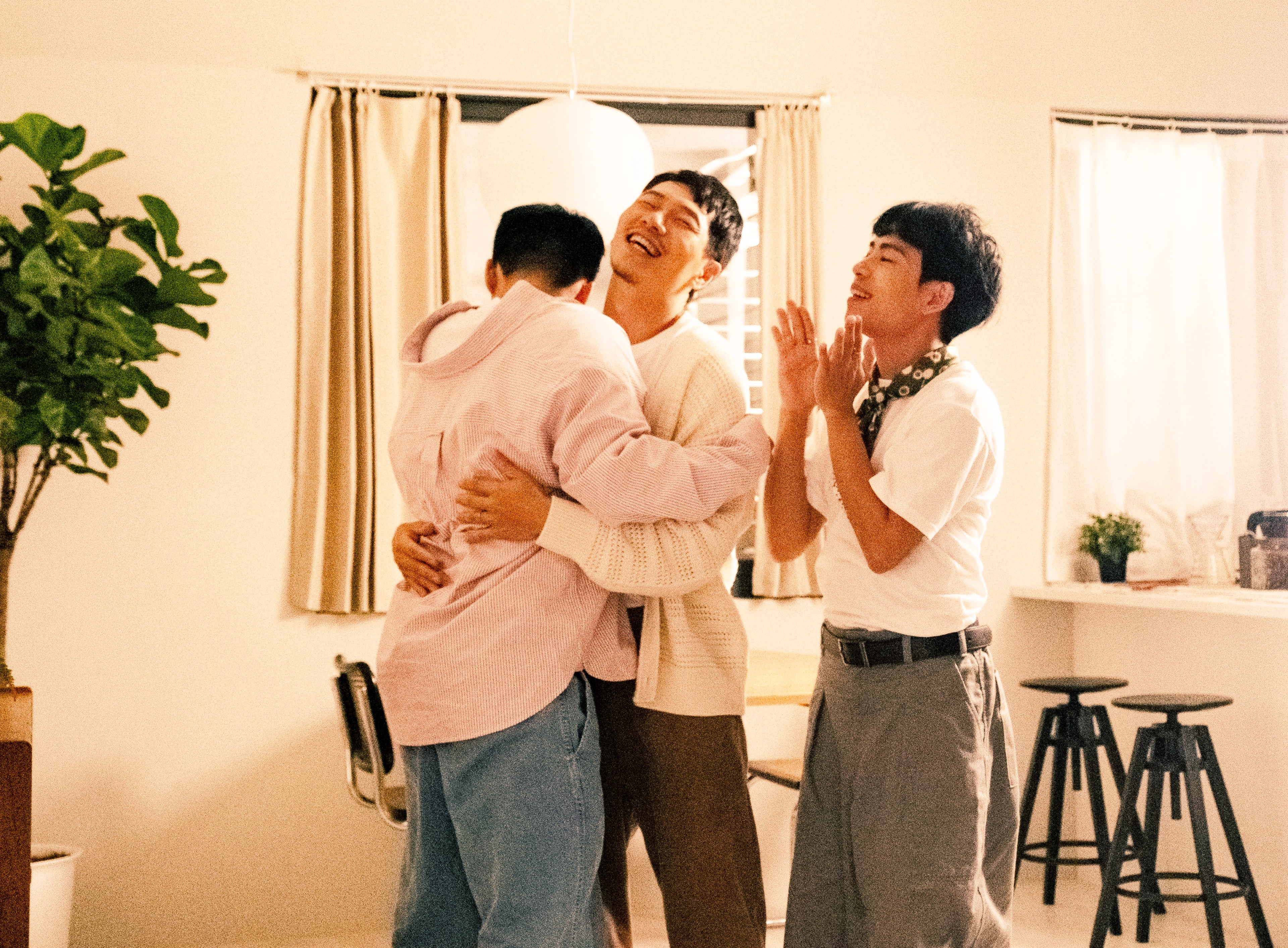 Three men celebrating together indoors, with two embracing and another clapping beside them.
