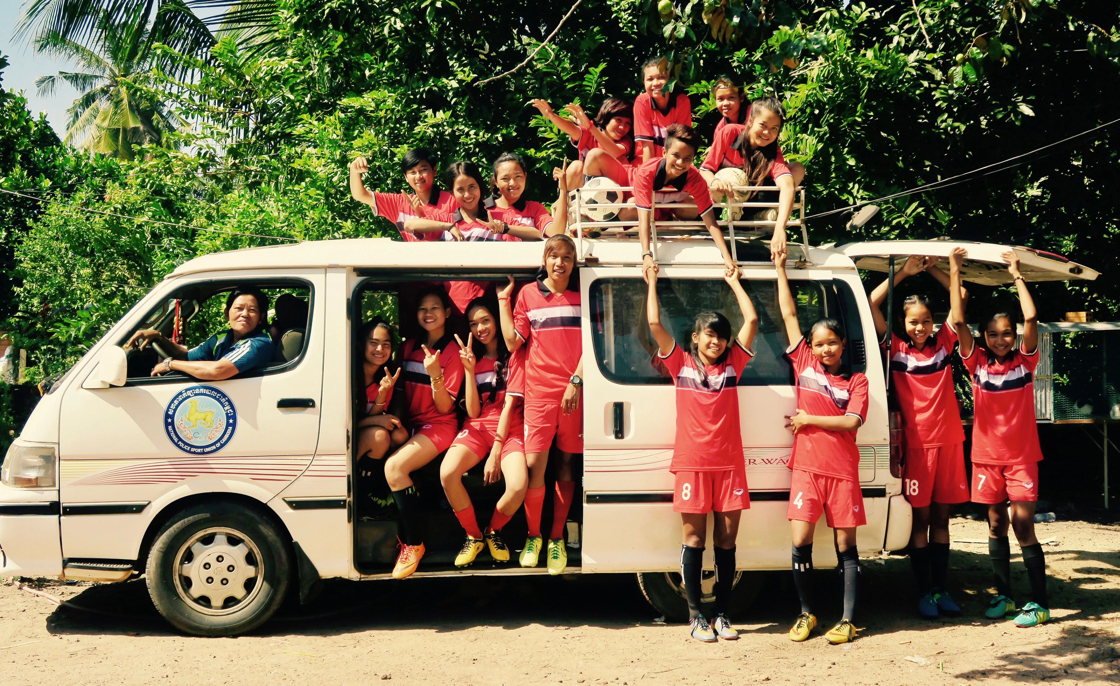 A women’s football team in red uniforms gathered around a white van, smiling and posing together in a tropical setting.