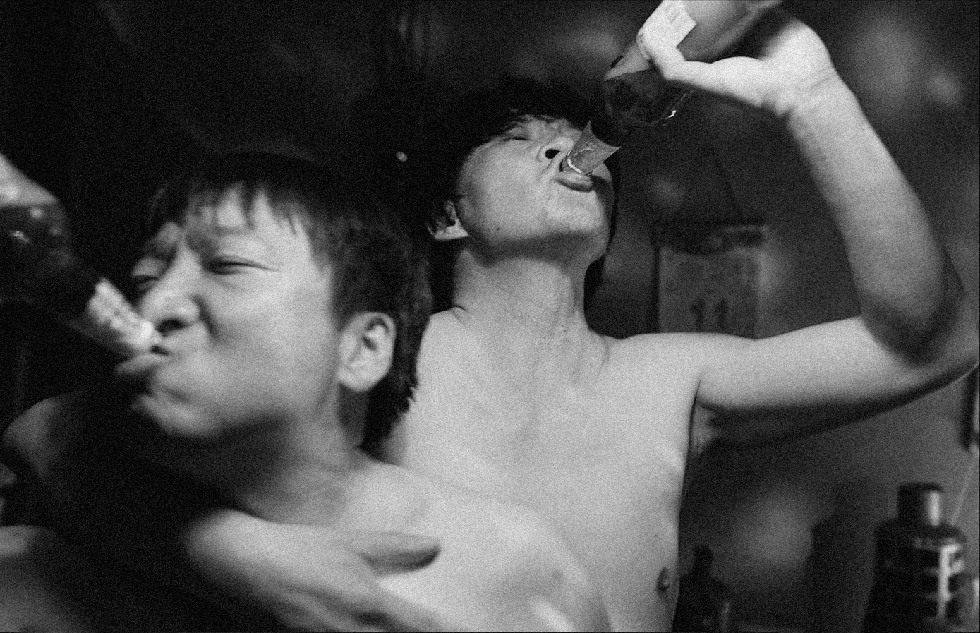 Two shirtless men drinking from bottles in a grainy black-and-white scene.