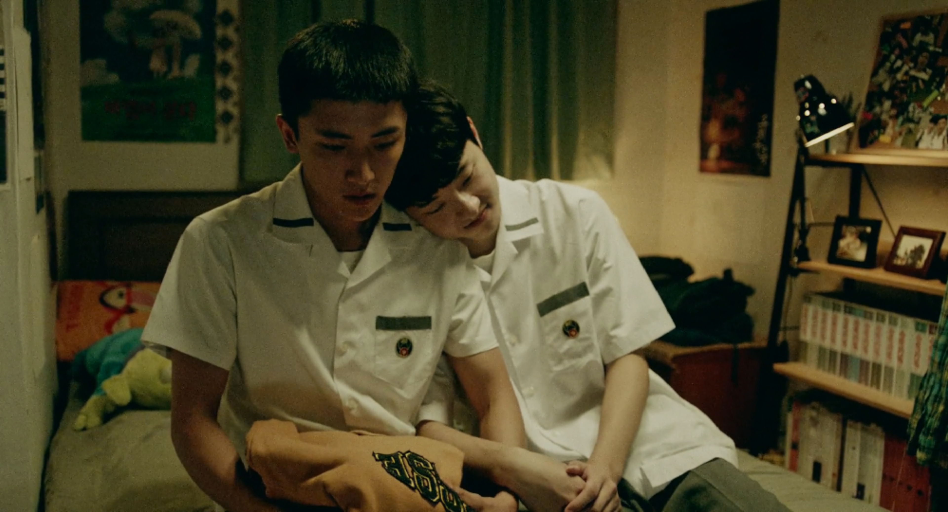 Two young men in school uniforms sitting closely together on a bed, one resting his head on the other’s shoulder.