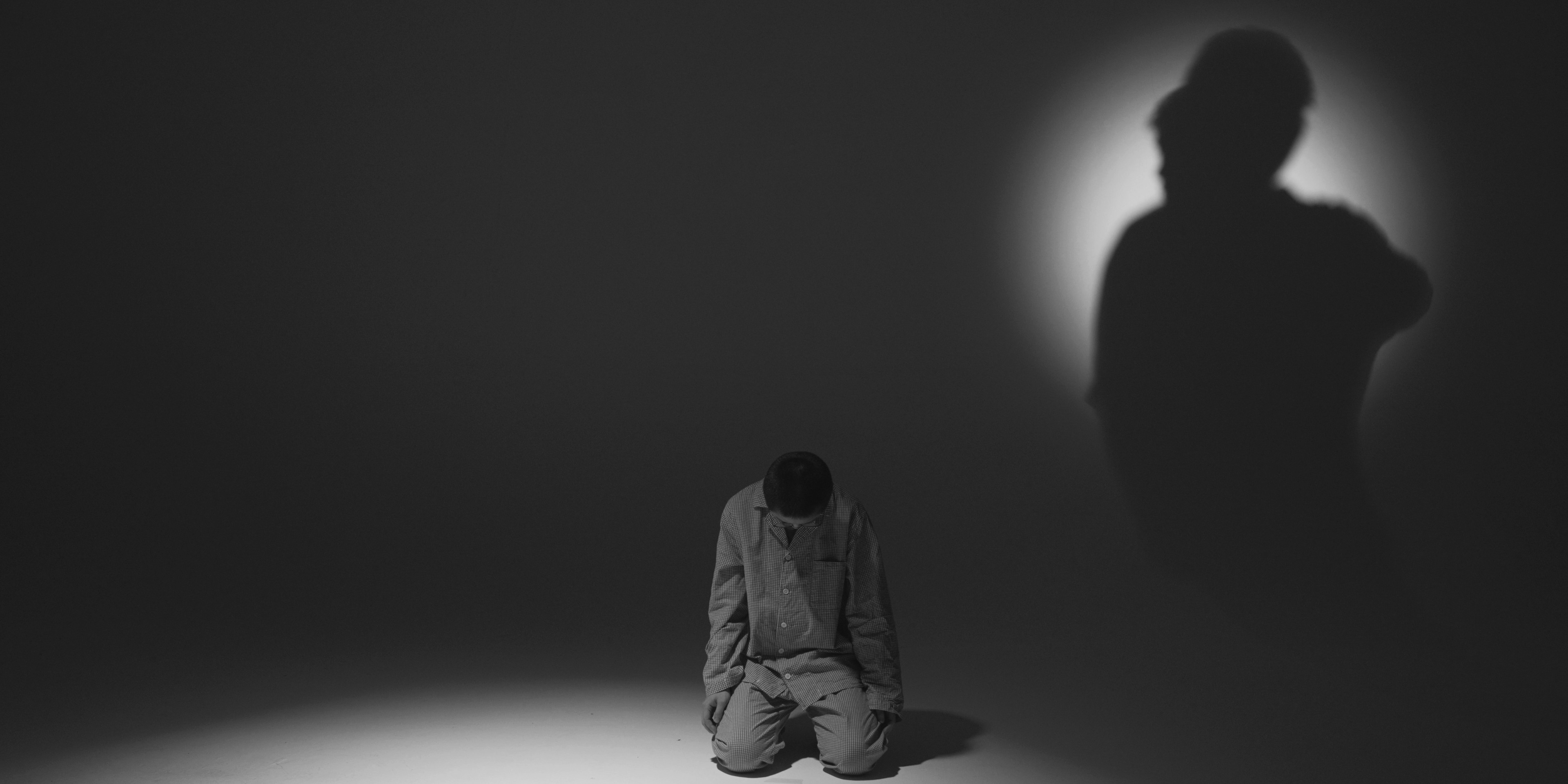 A person kneels with their head bowed in a dark space, while a large shadowy figure looms behind them against a circular light.