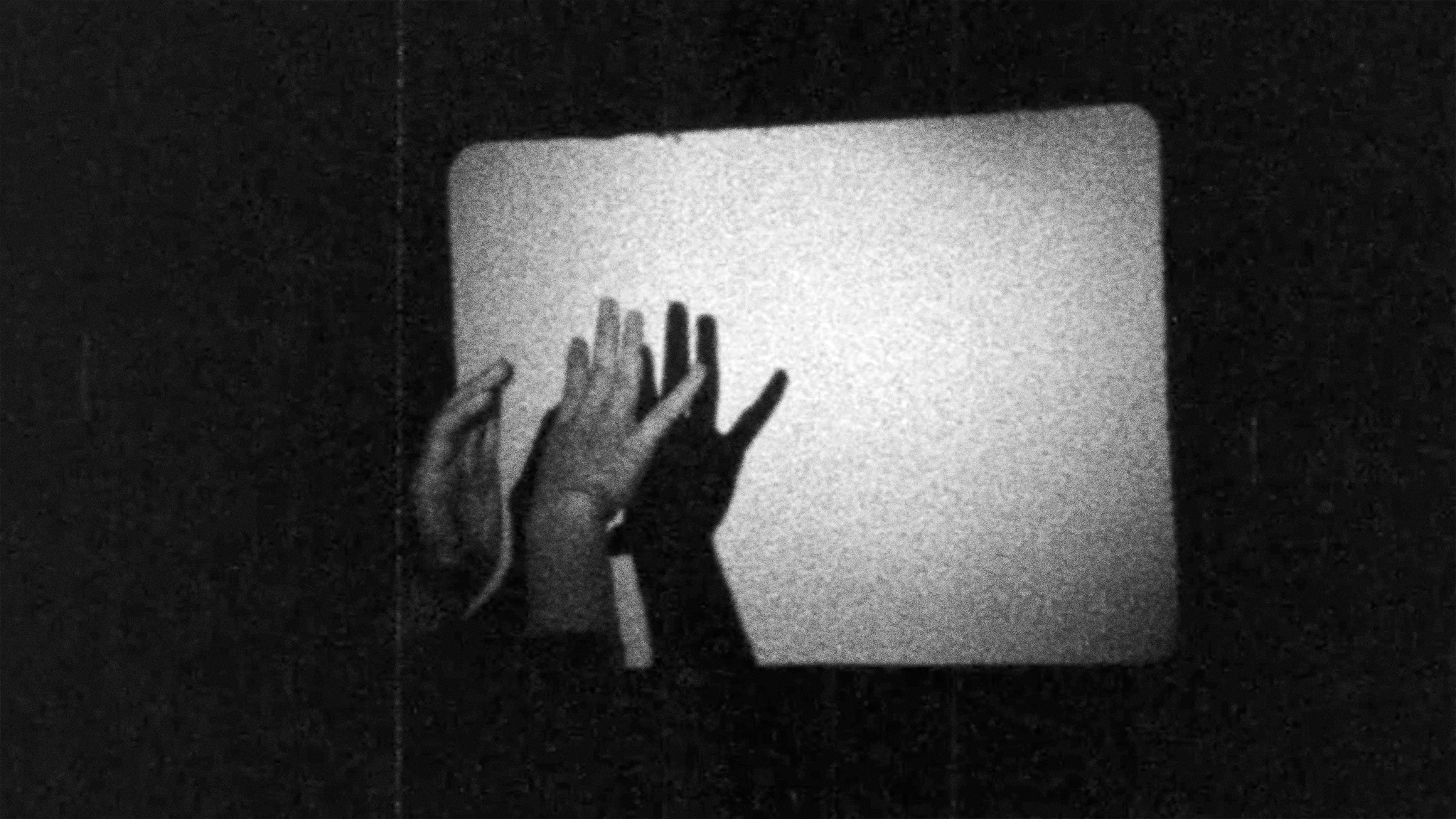 A grainy black-and-white image of several hands reaching toward a glowing rectangular screen.
