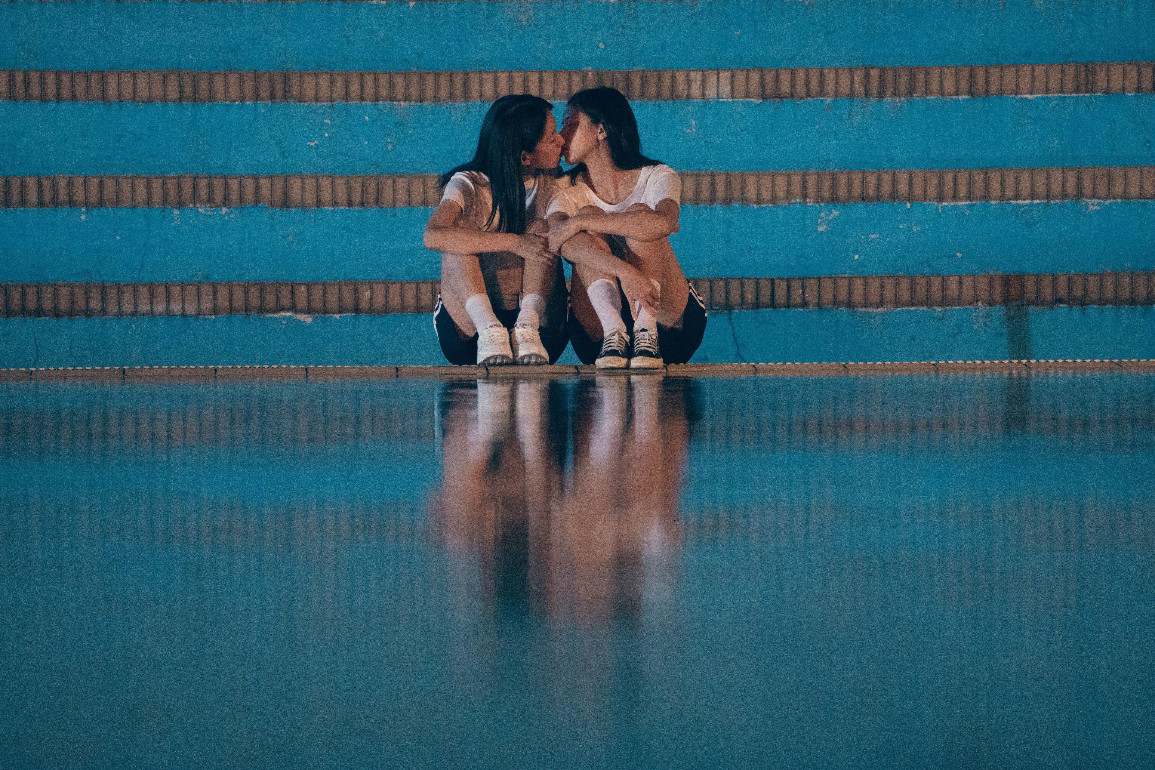 Two young women sitting close together on blue bleachers, kissing beside a reflective floor.