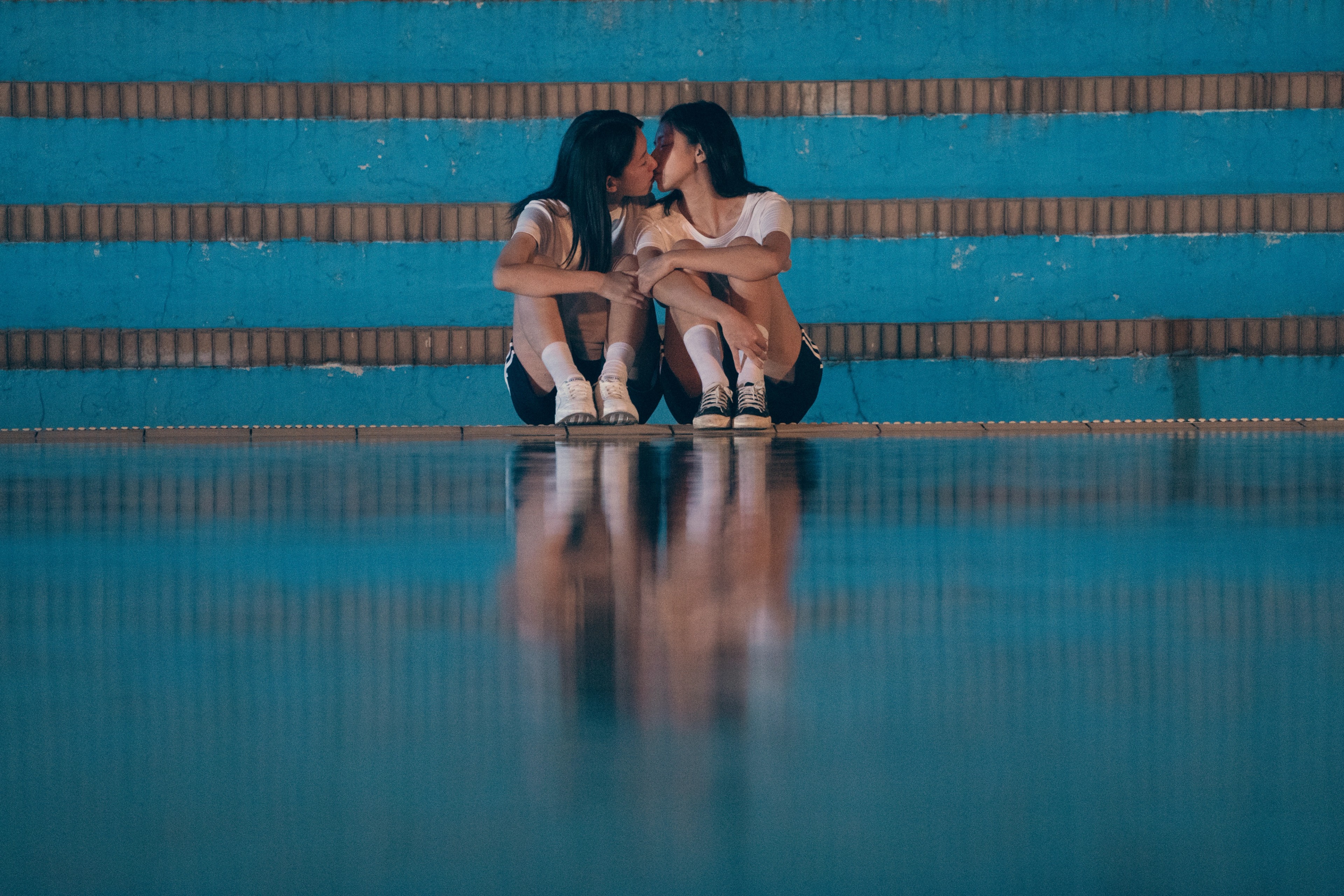 Two young women sitting close together on blue bleachers, kissing beside a reflective floor.