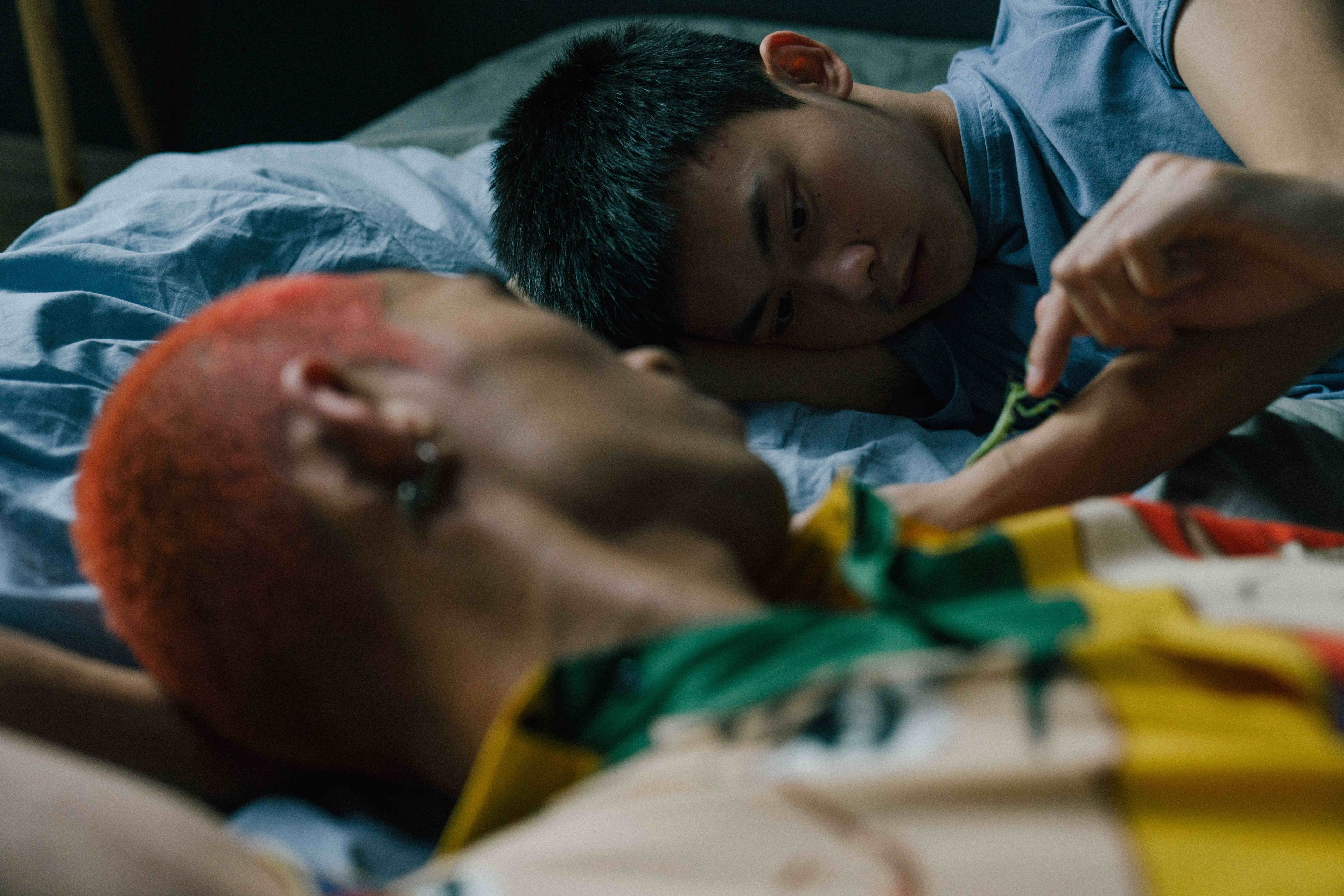 Two people lying closely together on a bed, one with bright orange hair while the other looks toward them.