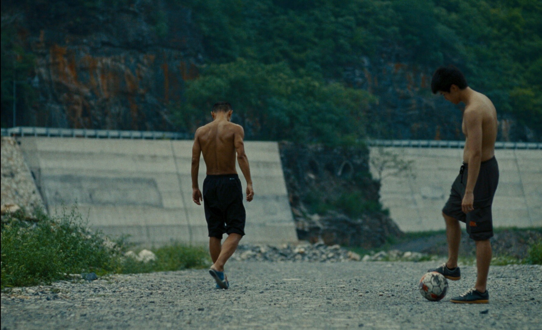 Two topless men kick a football next to a cliffside.