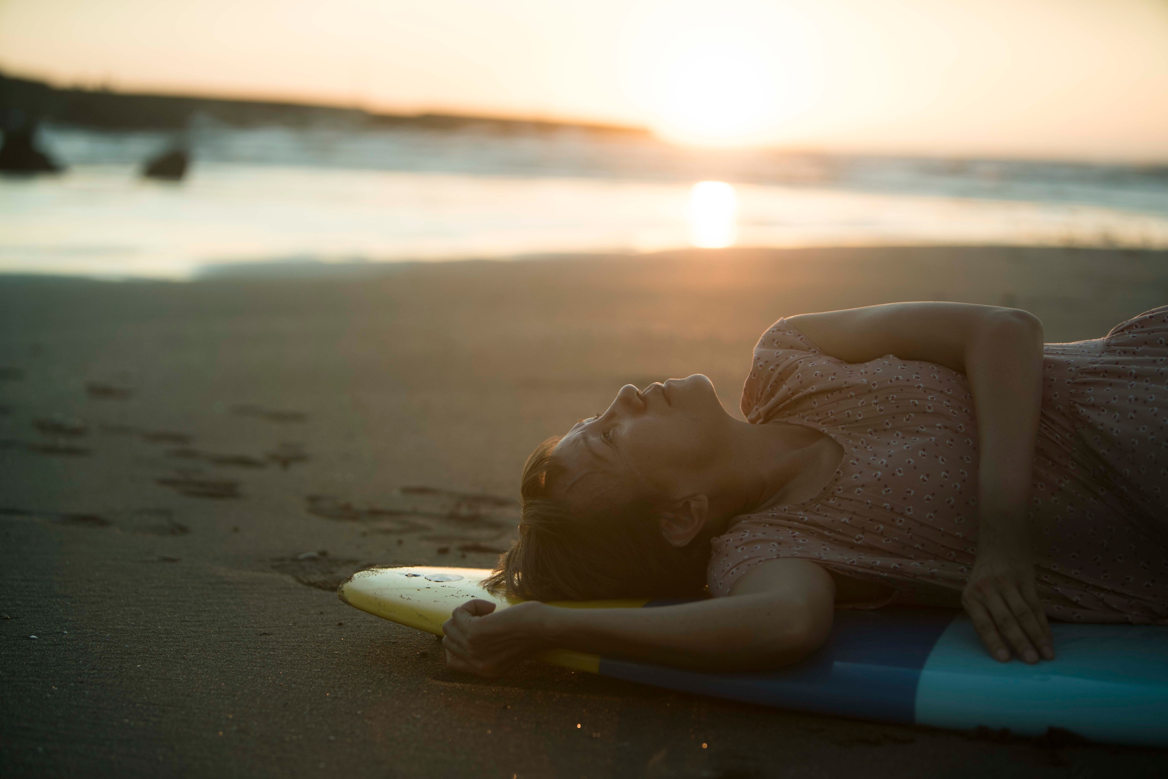 A woman lying on a surfboard on the beach at sunset, gazing upward beside the sea.