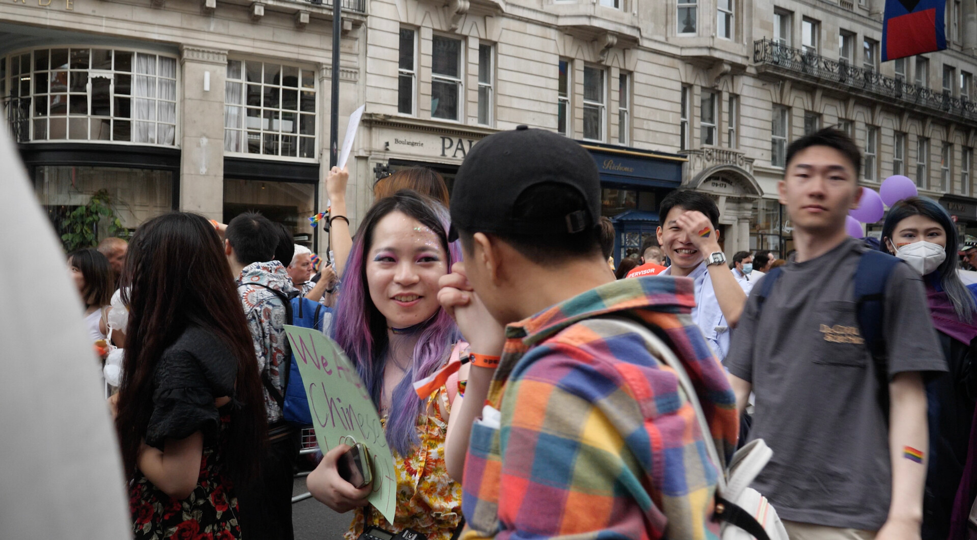 A crowd gathered at a Pride march on a city street, with several people holding signs and rainbow symbols.