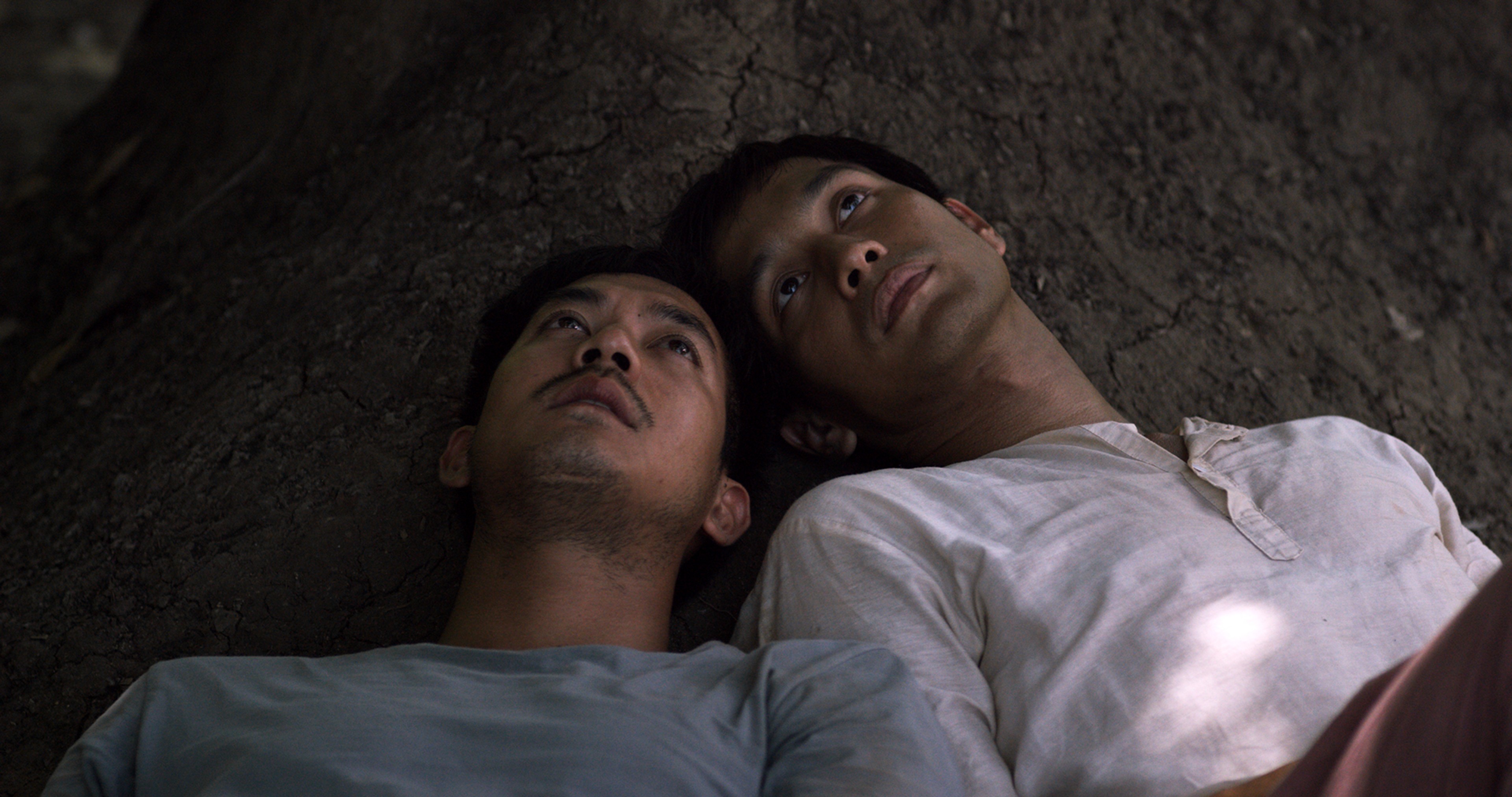 Two men lying closely side by side beneath a large tree, looking upward.