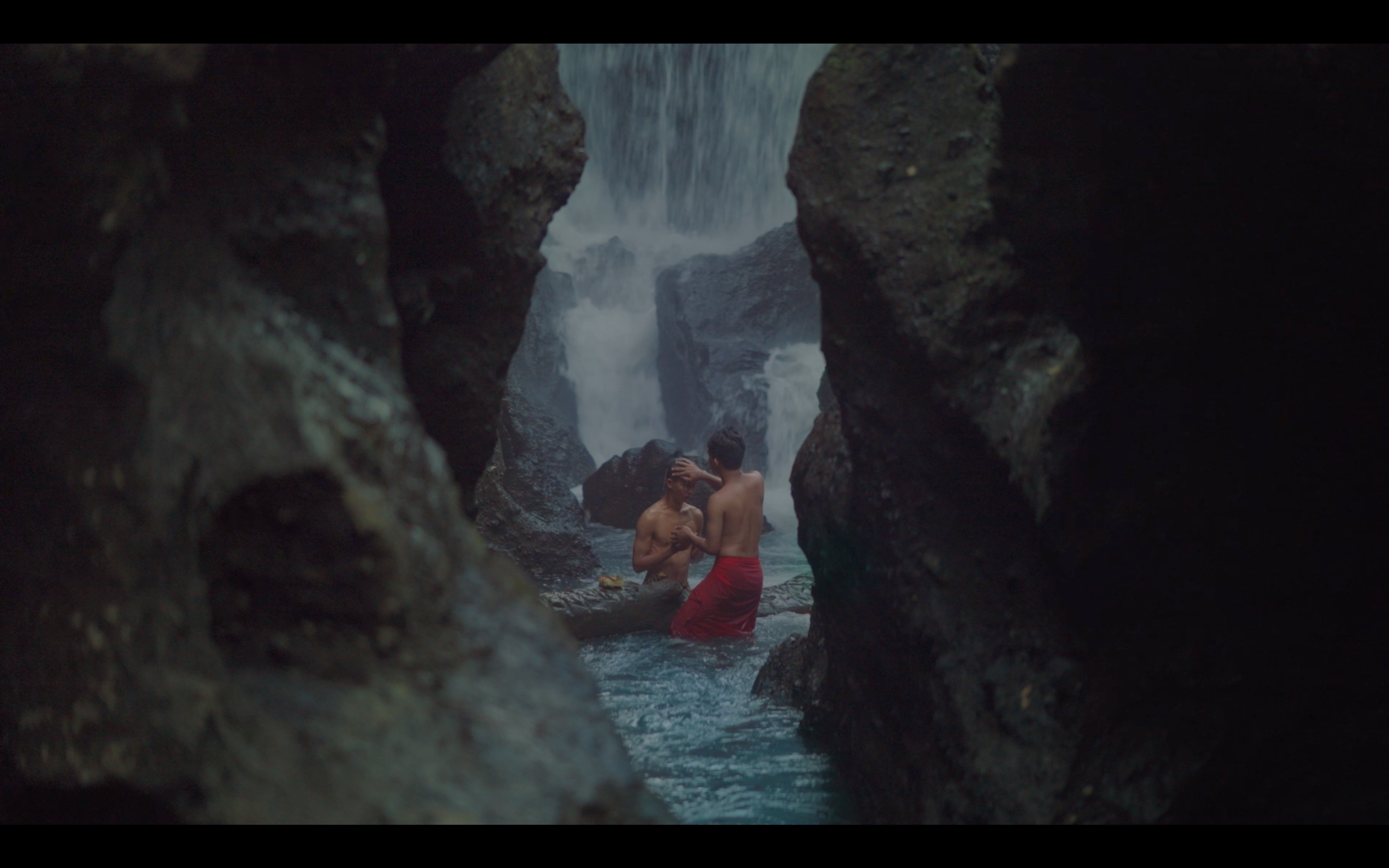 Two shirtless men standing closely together in a rocky pool beneath a waterfall.