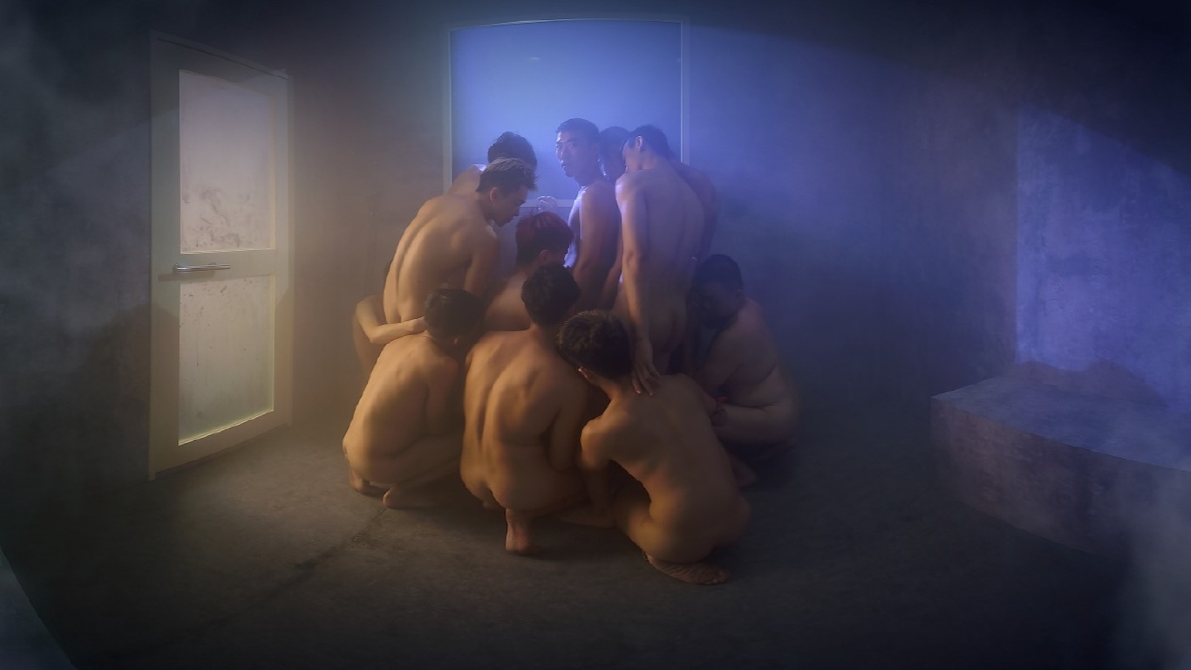 A group of nude figures gathered closely together in a dim, misty room illuminated by blue and warm light.