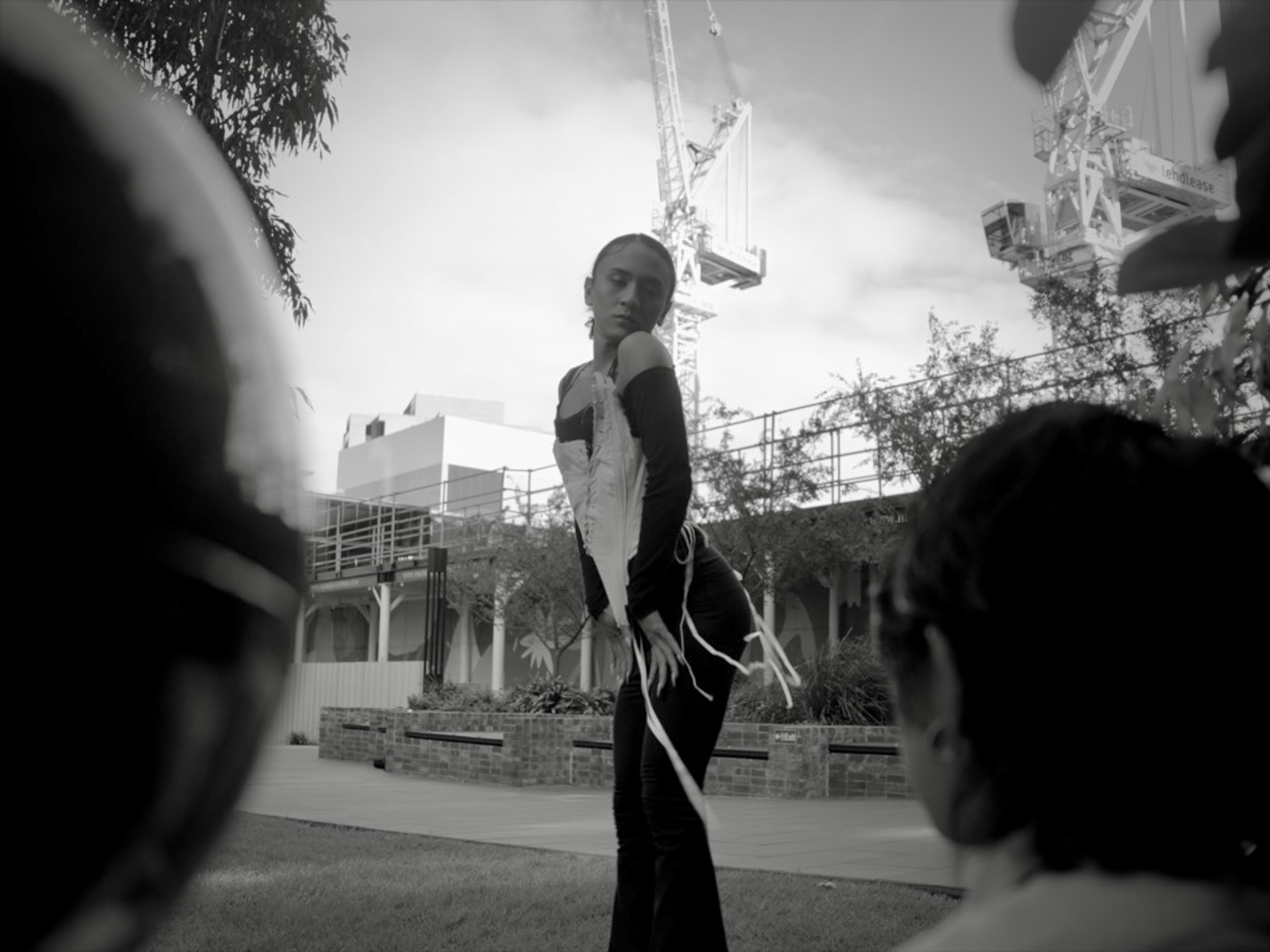 A performer standing outdoors in a black-and-white scene, framed by seated onlookers and construction cranes.
