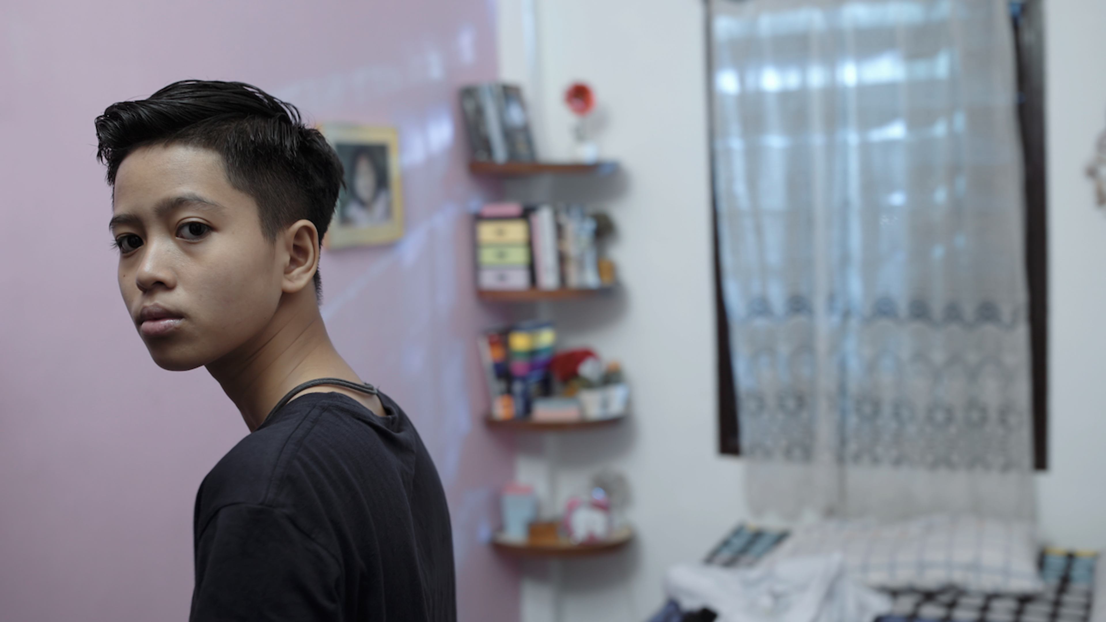 A young person with short hair looking back toward the camera in a softly lit bedroom.