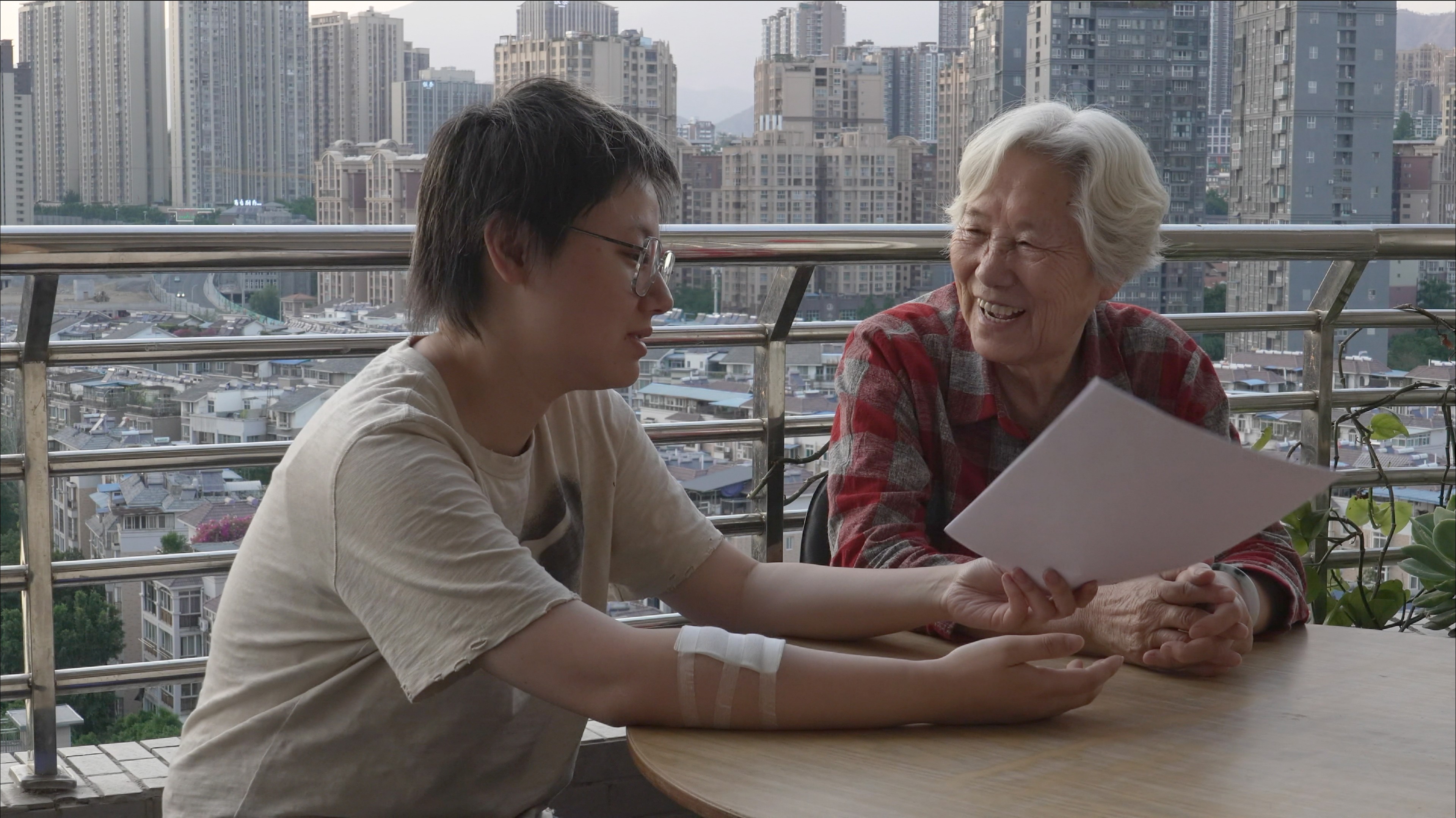 two figures sit at a table with skyscrapers in the backdrop.