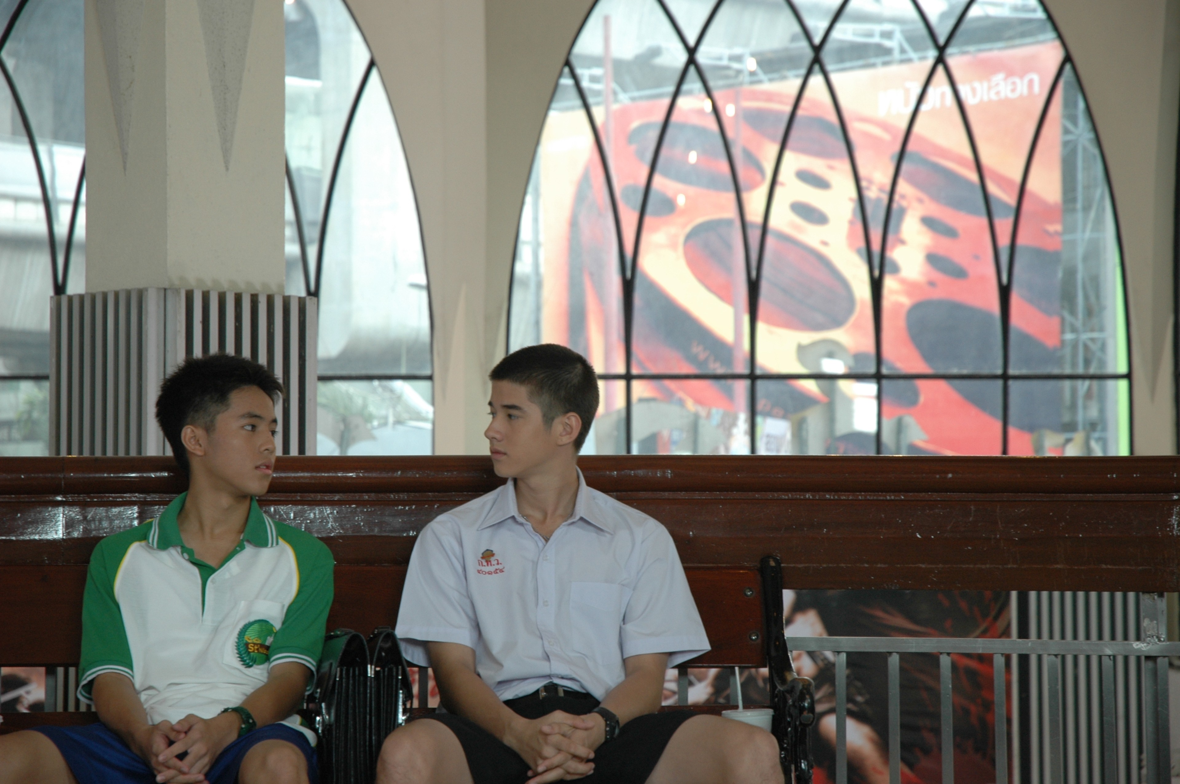 Two teenage boys in school uniforms sitting on a bench, looking toward each other.