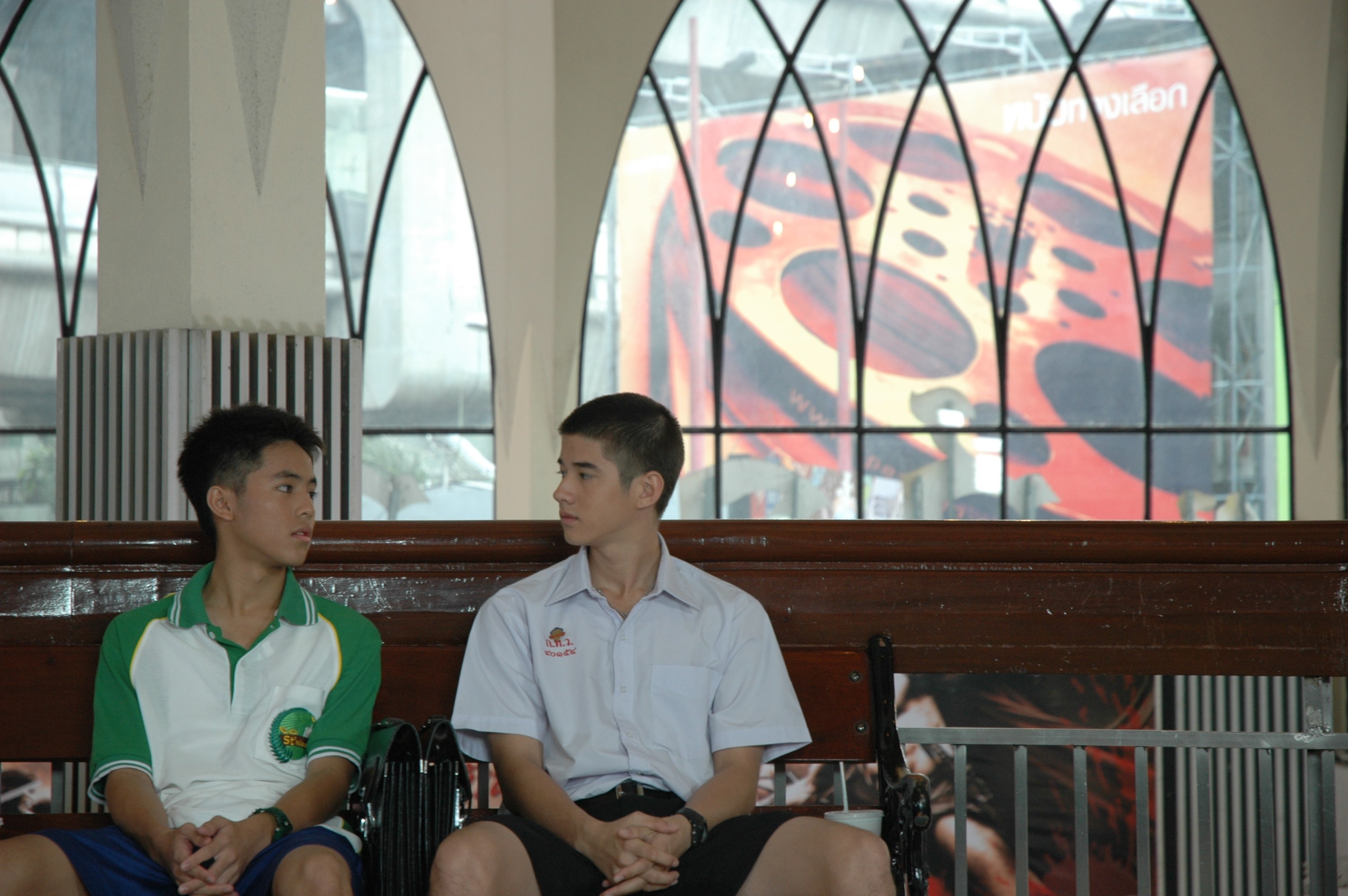 Two teenage boys in school uniforms sitting on a bench, looking toward each other.