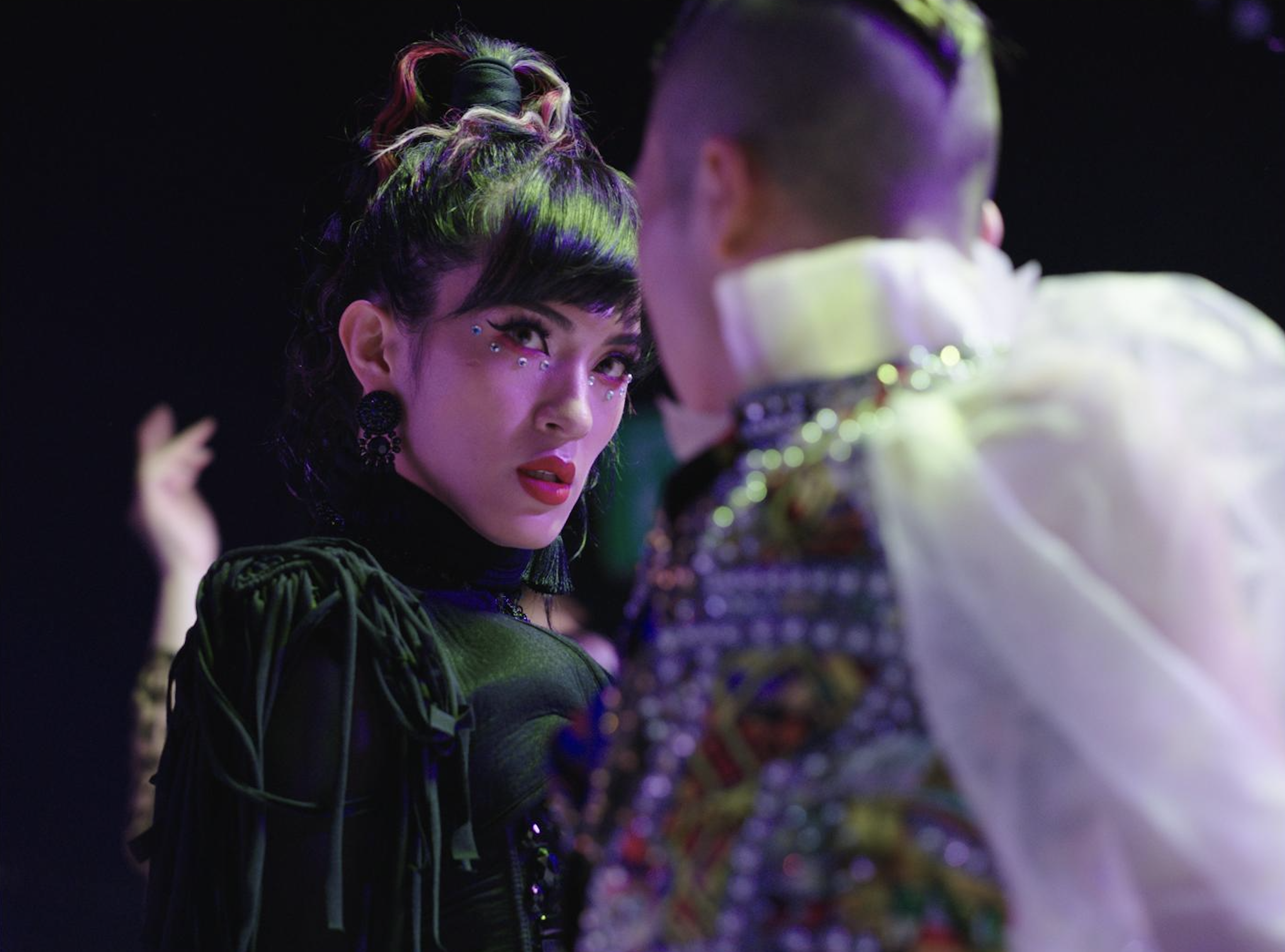 Two performers in elaborate costumes facing each other under purple stage lighting.