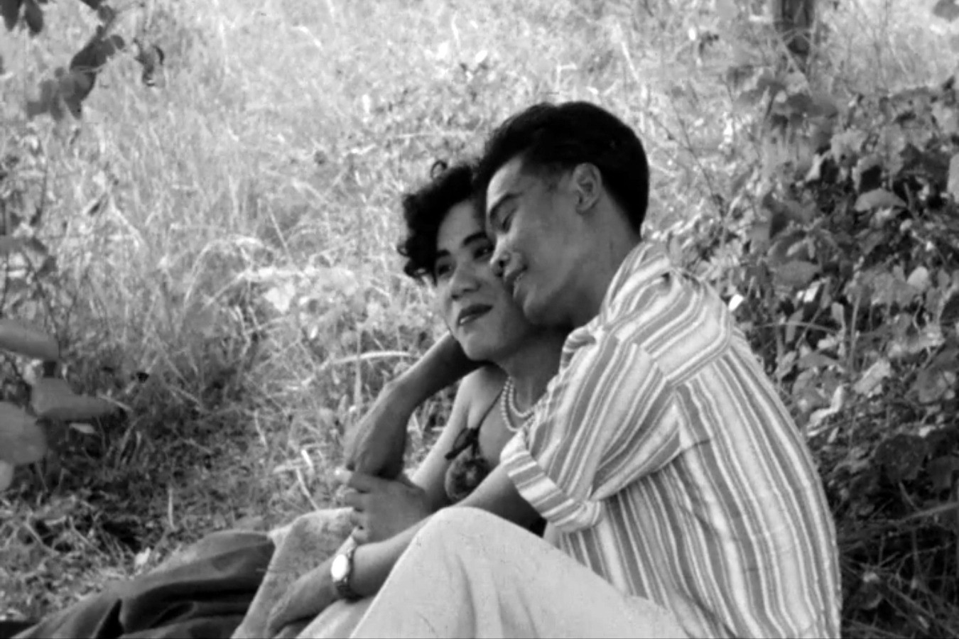Two people sitting closely together in tall grass, smiling and leaning toward one another in a black-and-white image.