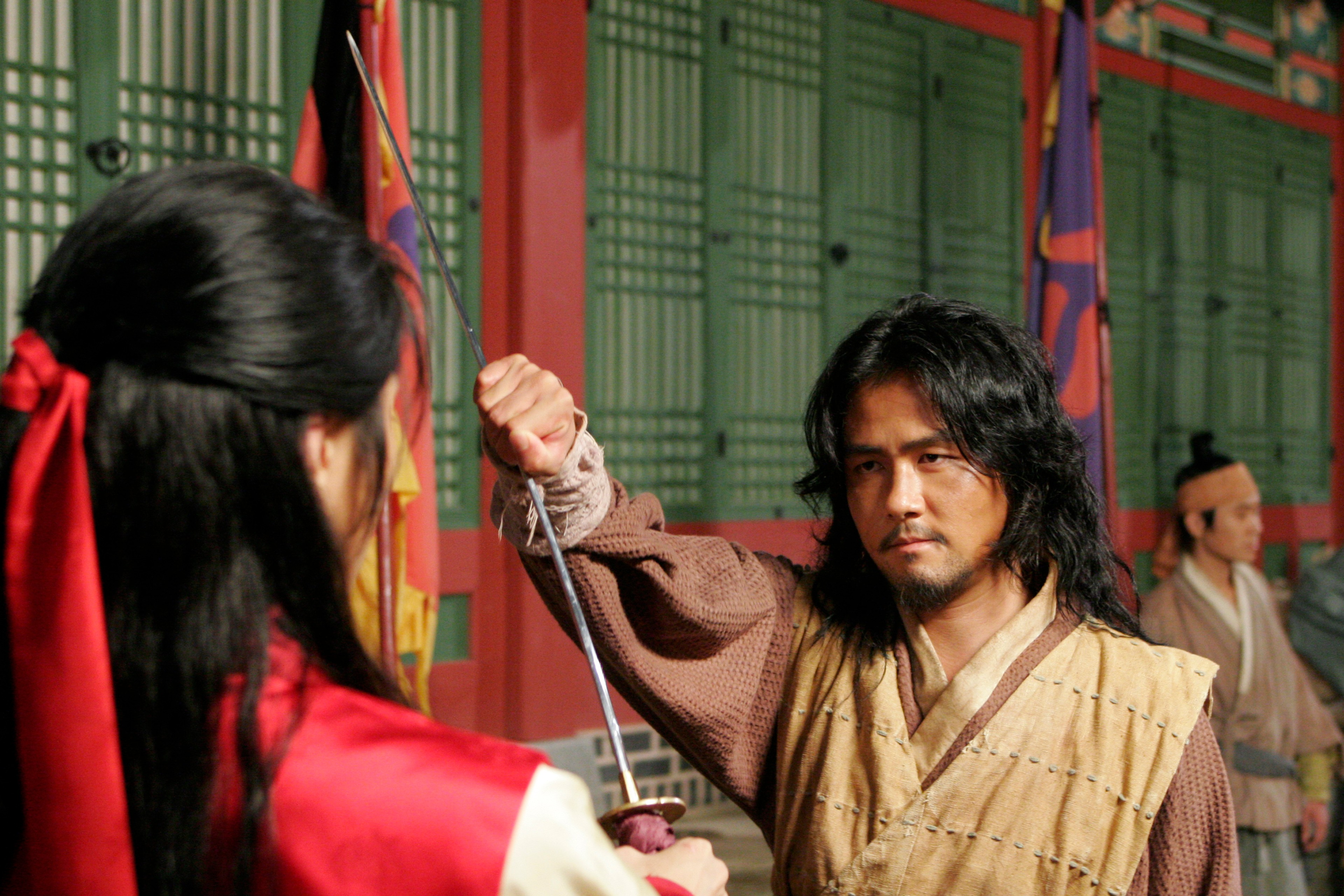 A man in historical dress holding up a sword toward a person in red, outside a traditional building.